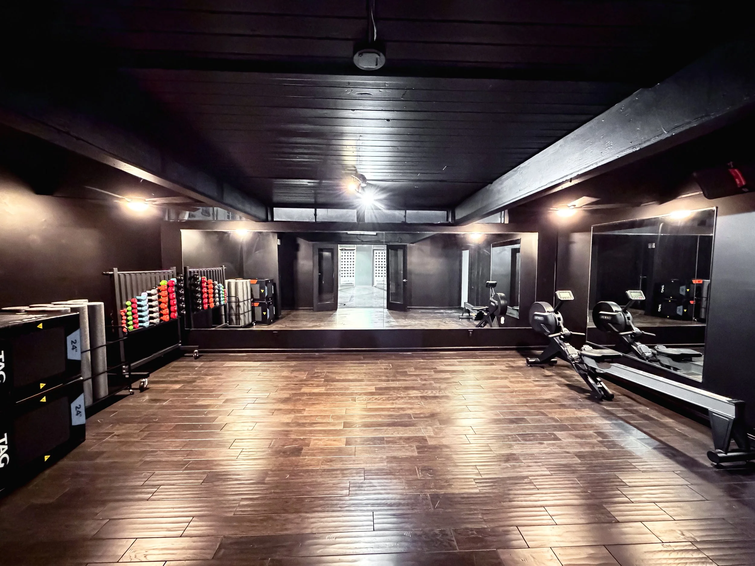 Empty black-walled fitness studio with wooden flooring, mirrored wall, gym equipment, and a workout area with rowing machines.