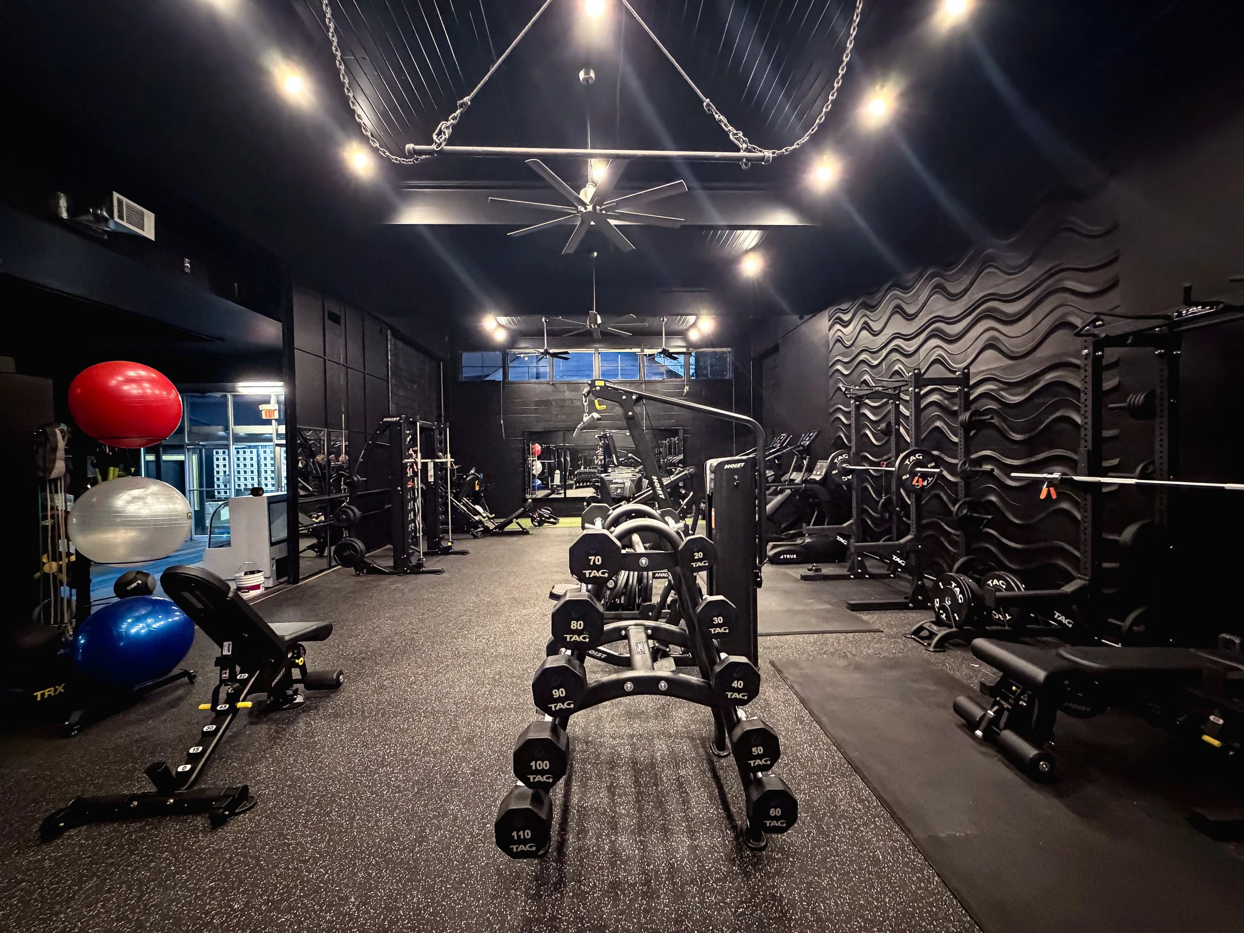 Empty gym with various workout equipment, including dumbbells, exercise balls, weight benches, and machines, with dark walls and ceiling, and a large mirror reflective of the gym's interior.