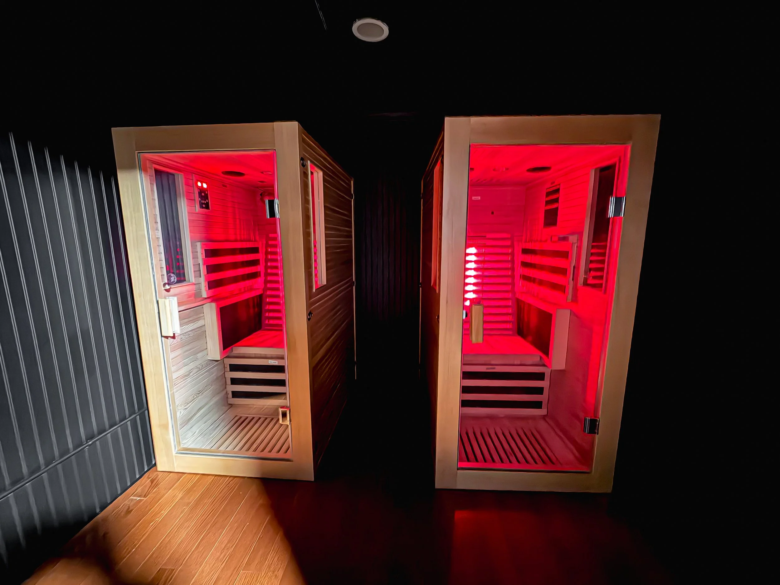 Two infrared saunas with glass doors and wooden interiors illuminated with red light, placed side by side in a dark room.