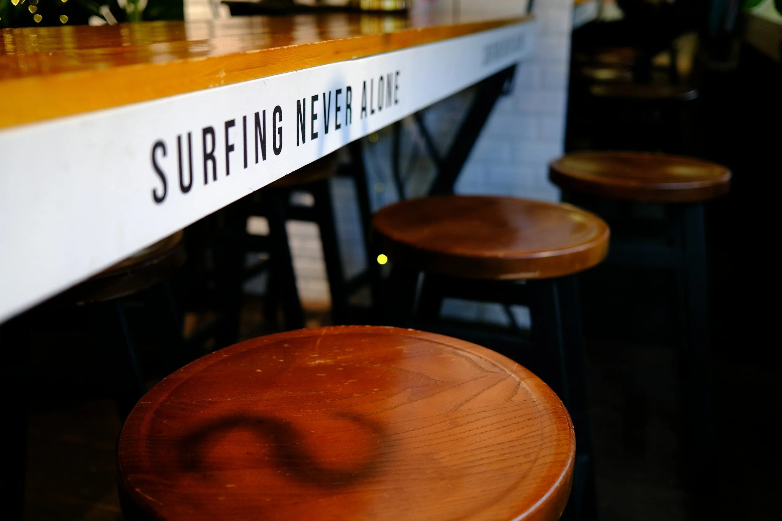 Wooden barstools lined up along a counter with a sign that says 'Surfing Never Alone' in a cafe or bar.