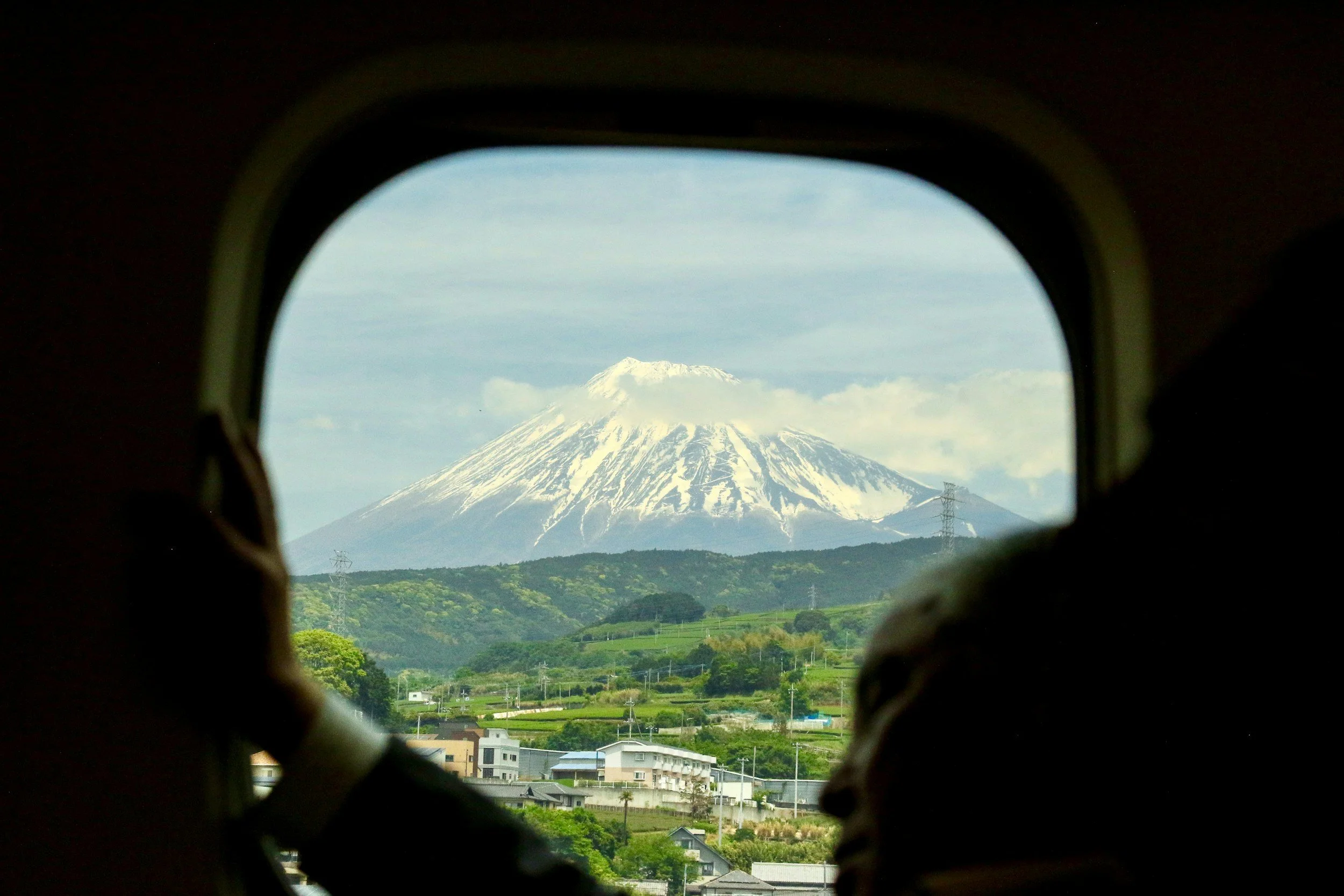 View of a snow-capped mountain, Mount Fuji, through an airplane window, with landscapes and buildings in the foreground.