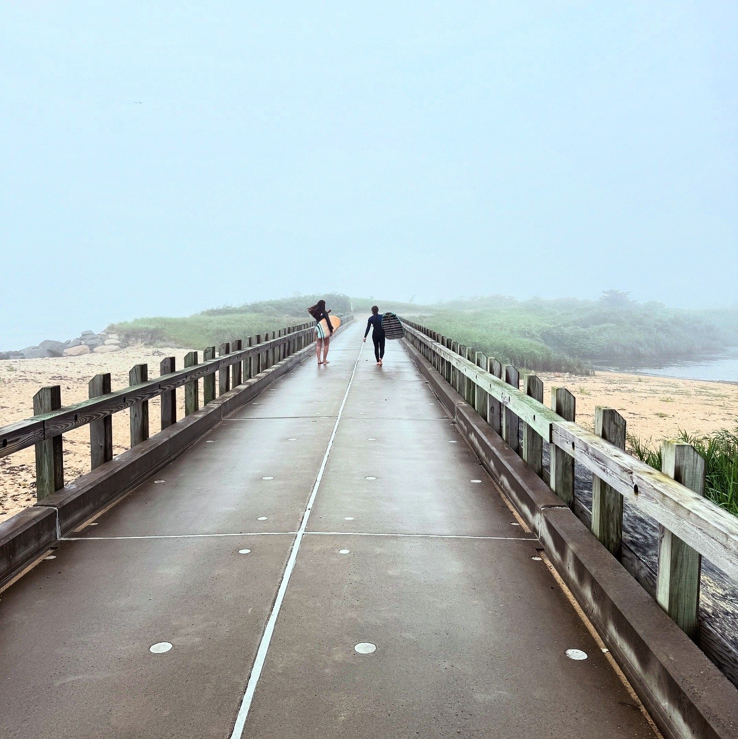 A foggy concrete pier with wooden railings on both sides, leading towards the foggy horizon. There are two people walking away from the camera, one carrying a surfboard and the other carrying a bag, with sand and greenery on either side.