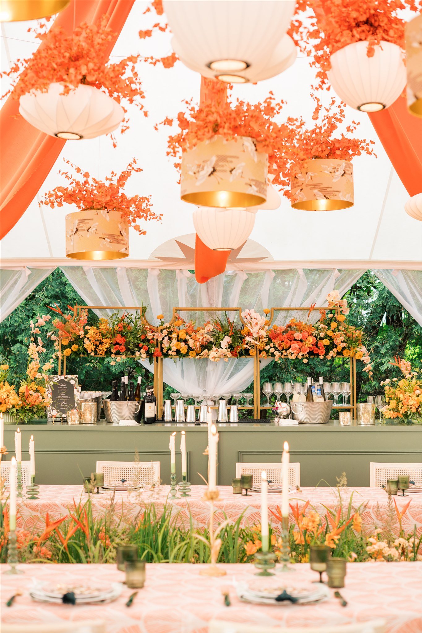 Decorated event tent with hanging paper lanterns, floral arrangements, and a bar with drinks and glassware.