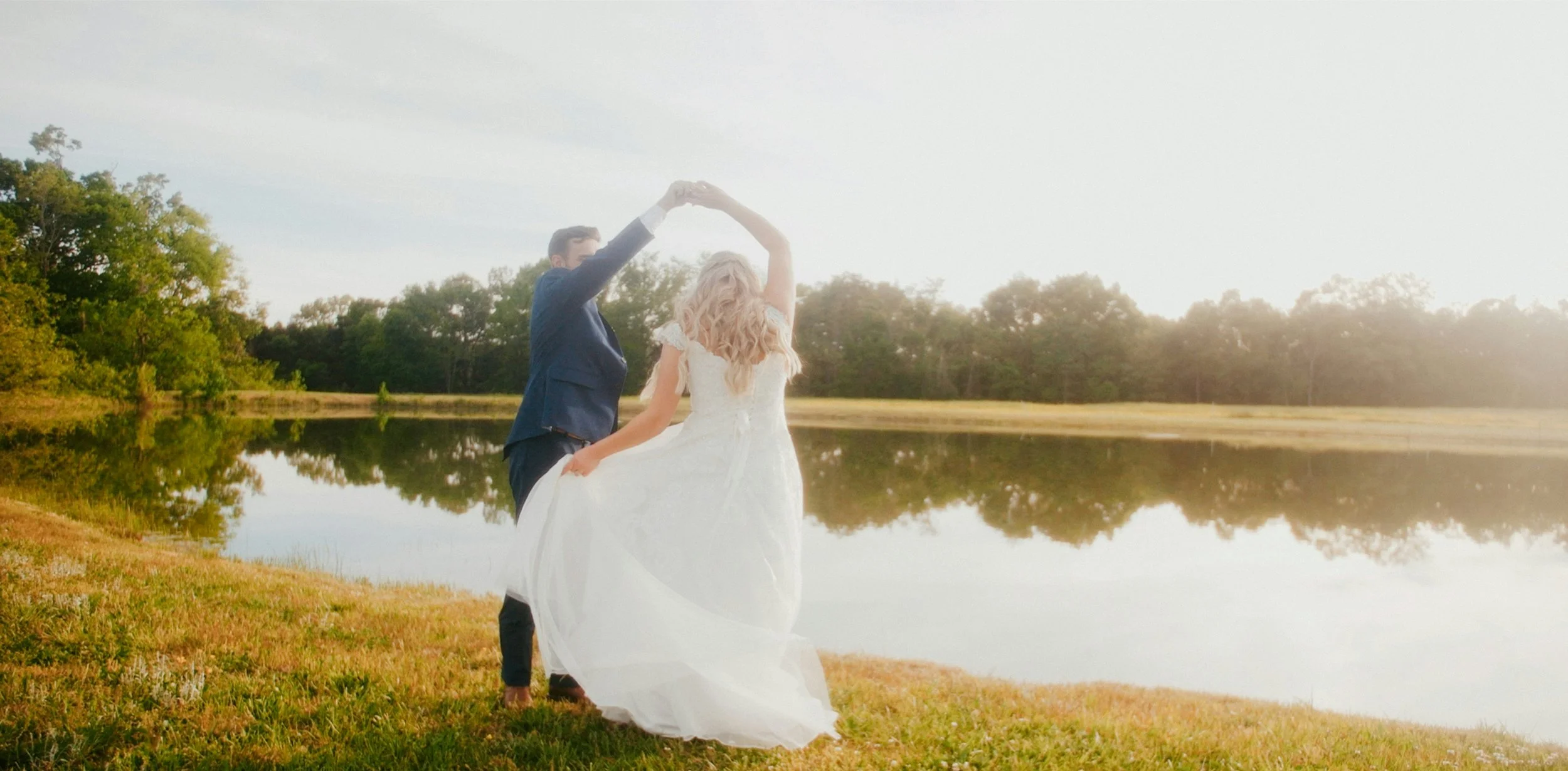 A couple in wedding attire dancing by a pond during sunset with trees in the background.