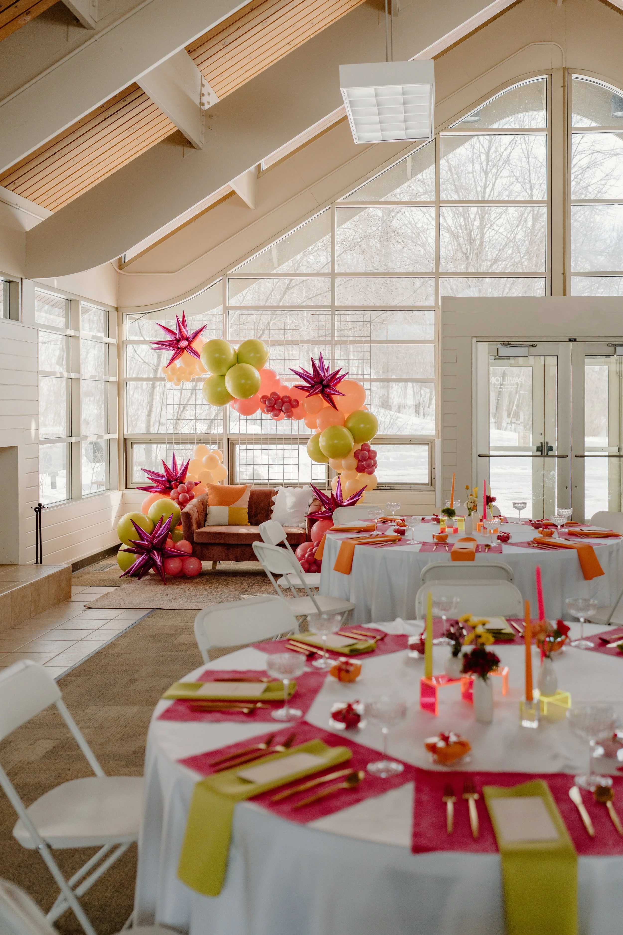 Decorated indoor party space with round tables covered in white tablecloths, set with colored napkins, utensils, and glassware. A backdrop of colorful balloons and star-shaped decorations surrounds a cozy brown sofa with pillows. The room has large windows and high, angled ceilings with wooden beams and a hanging light fixture.