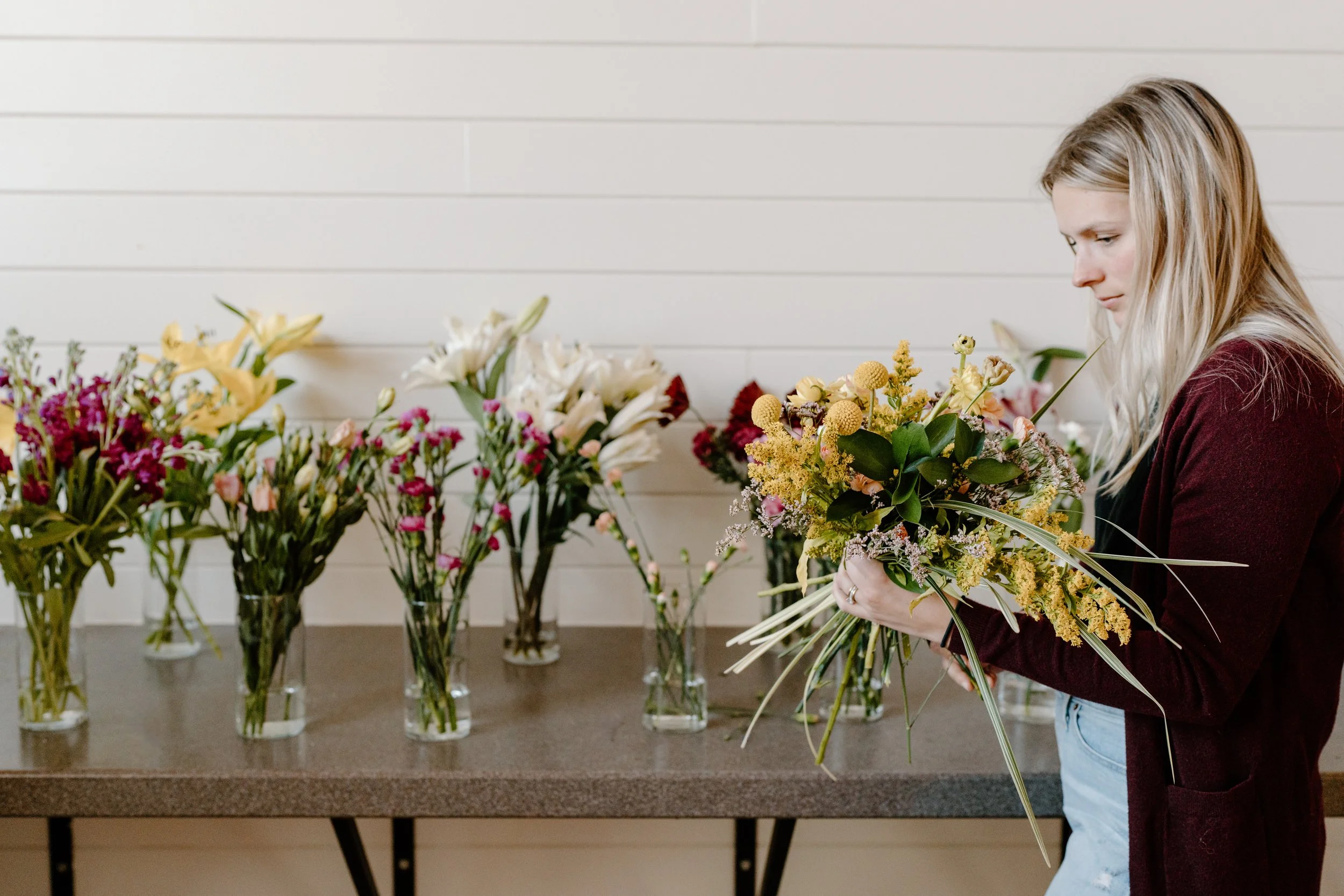 A woman arranging a bouquet of yellow and green flowers indoors with a row of various flower arrangements in vases on a table behind her.