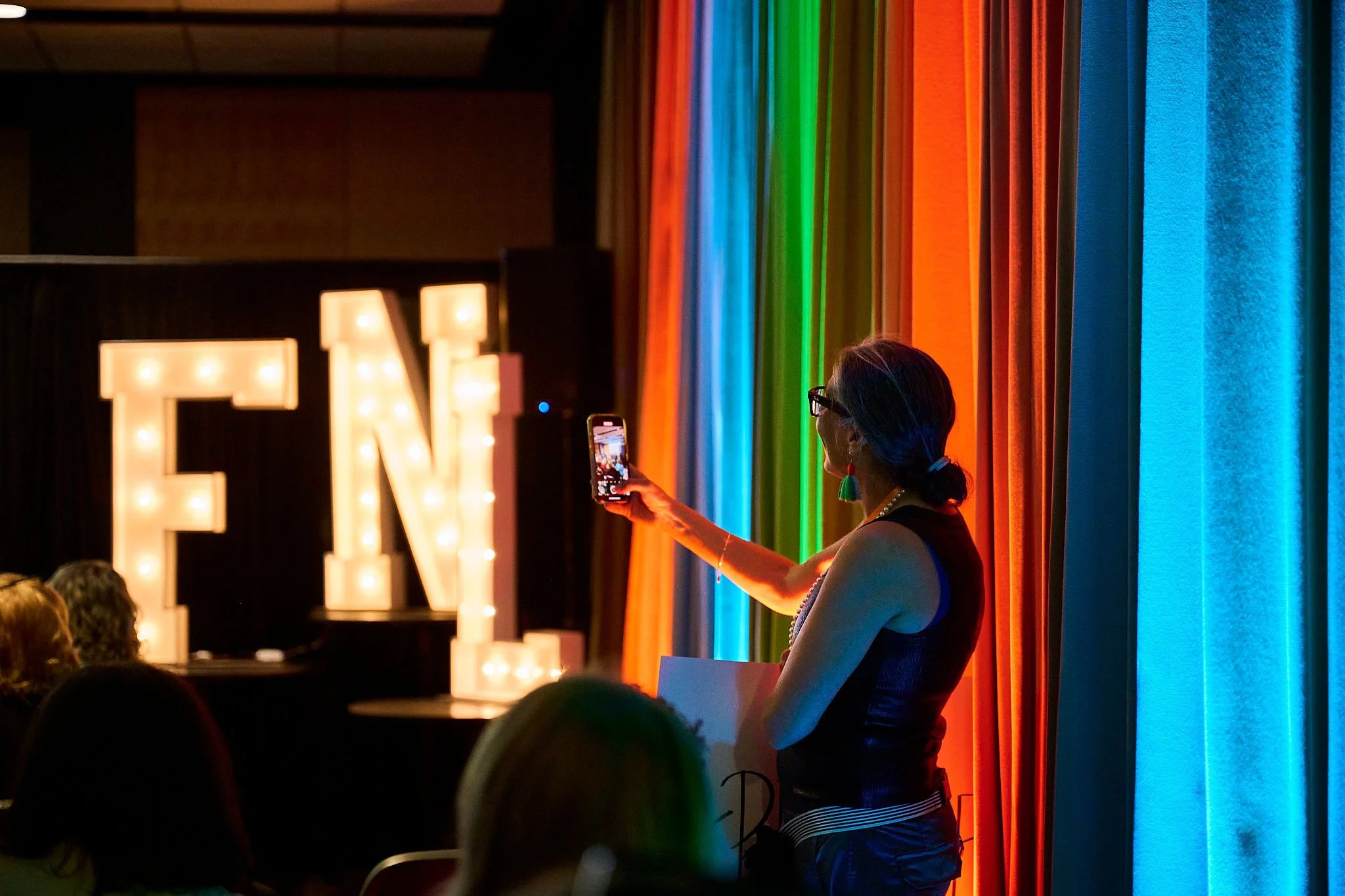 A woman taking a photo with her phone at a pride event, with a large illuminated letter 'E' and colorful rainbow curtains in the background.