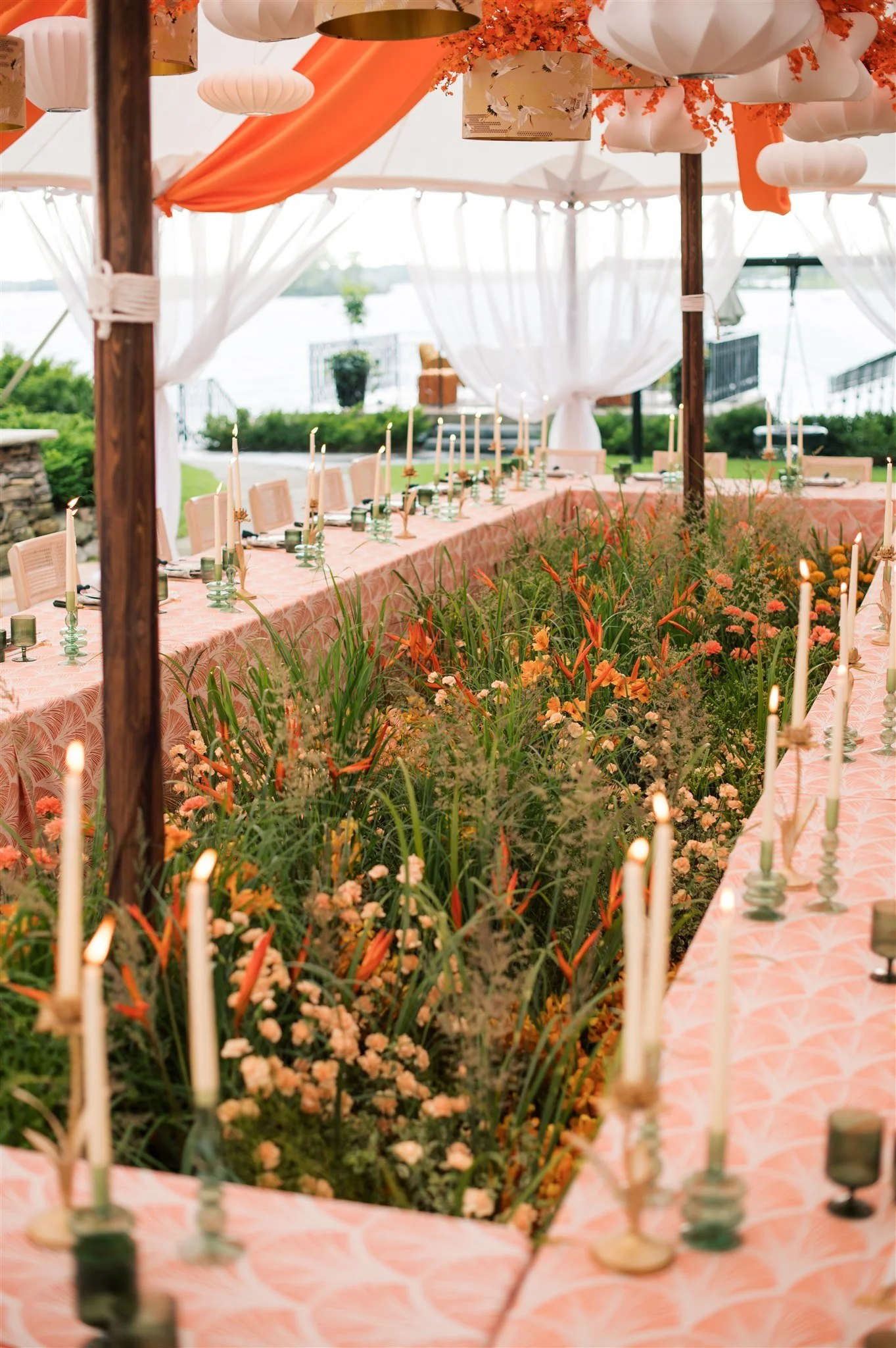 A decorated outdoor dining area with a long table set with white candles, surrounded by lush orange and pink flowers, under a canopy with orange drapes and lanterns, near a body of water.