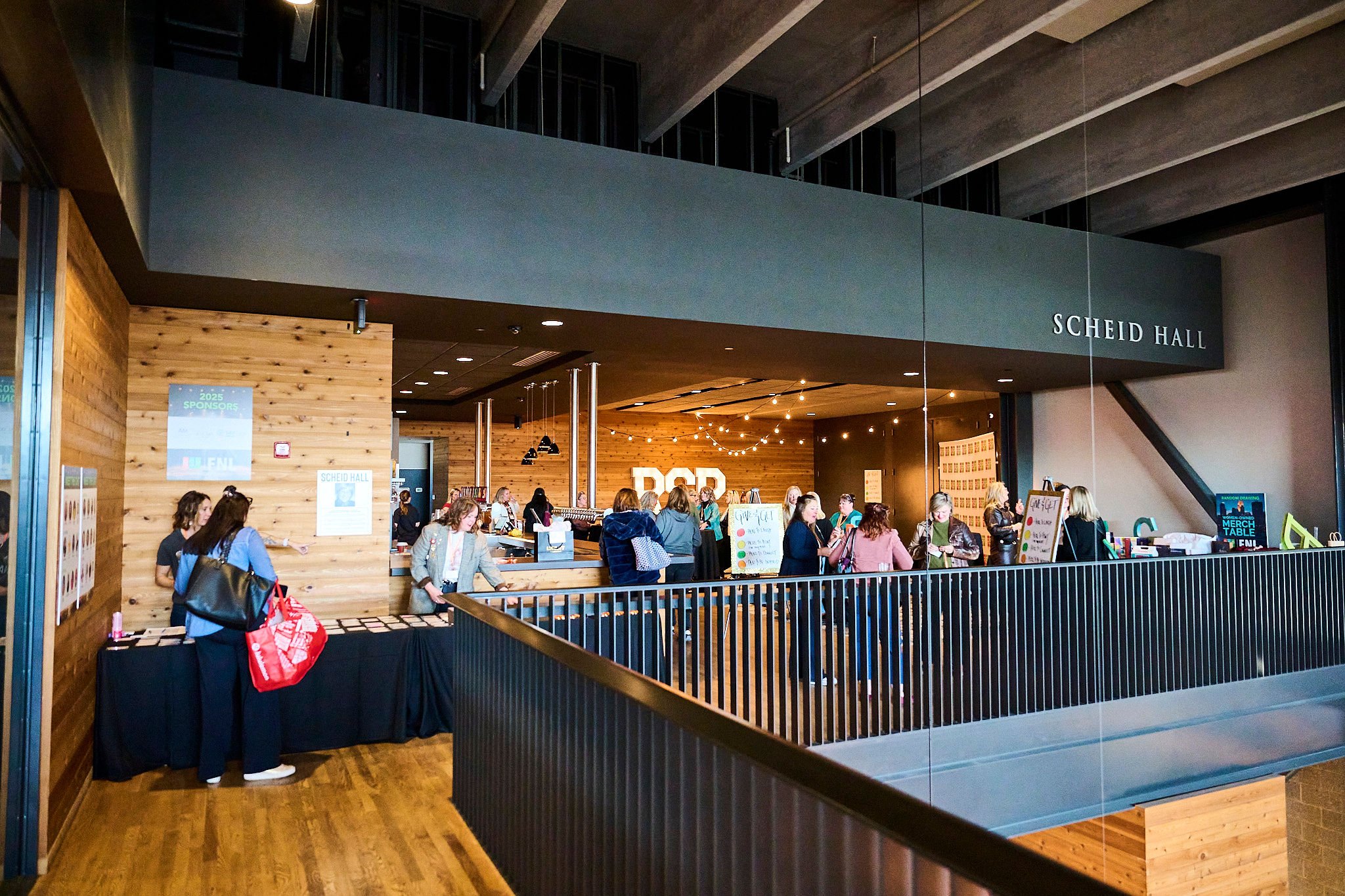 People lined up at a registration or check-in area inside a modern building with wood-paneled walls and a sign reading 'Scheid Hall'.