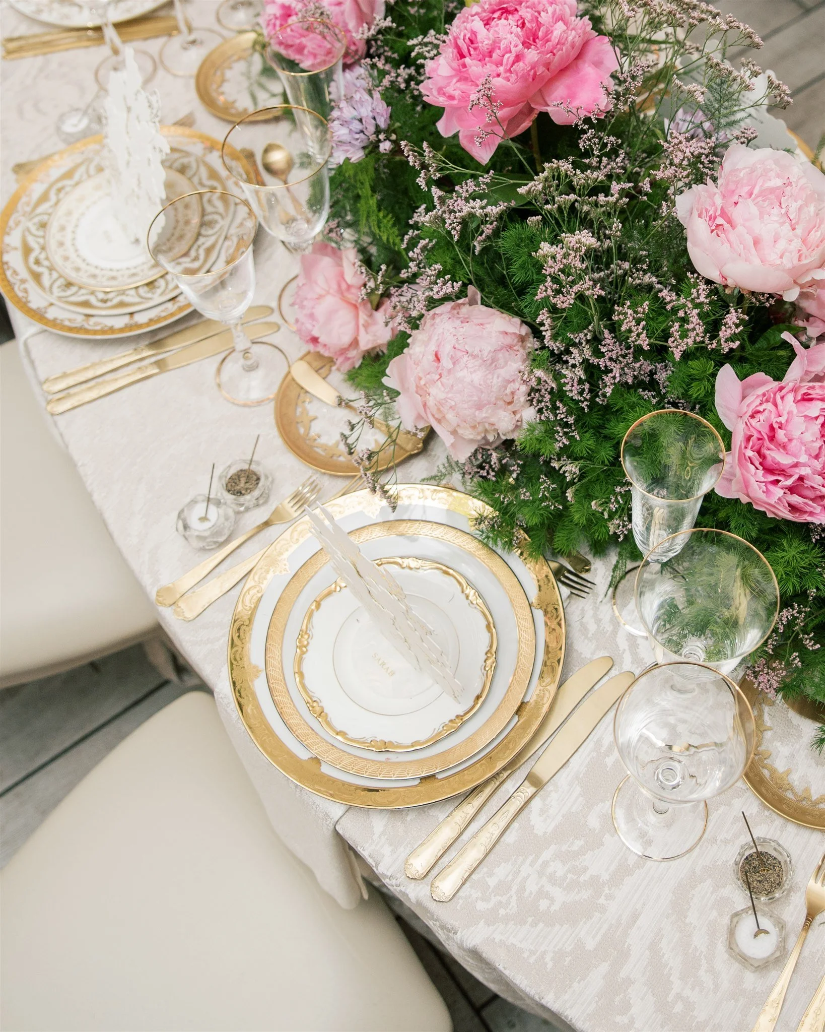Elegant table setting with gold-rimmed white china plates, gold flatware, clear glass stemware, and a floral centerpiece of pink peonies, lavender, and greenery on a white tablecloth.