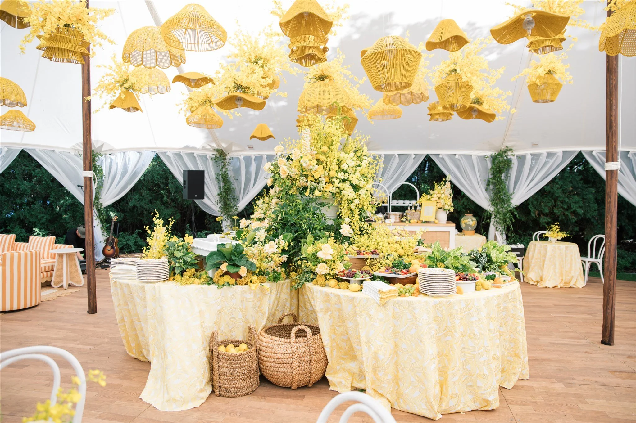 Decorated outdoor event tent with yellow and white floral arrangements, hanging yellow lampshades, and a table with food, plates, baskets, and desserts. Green foliage and trees are visible outside the tent.