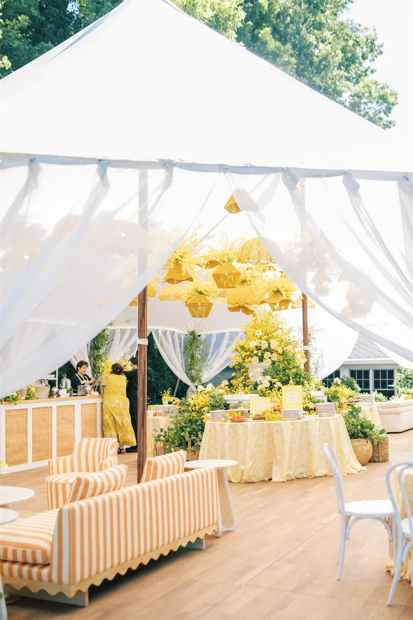 Bright outdoor event area decorated with yellow floral arrangements, curtain drapes, and hanging lanterns. There are tables with yellow tablecloths, striped yellow and white chairs, and a buffet station in the background.