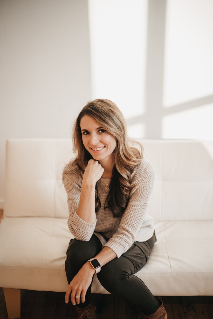 Woman sitting on a white sofa, smiling, with her hand near her chin, in a bright room with sunlight coming through windows.