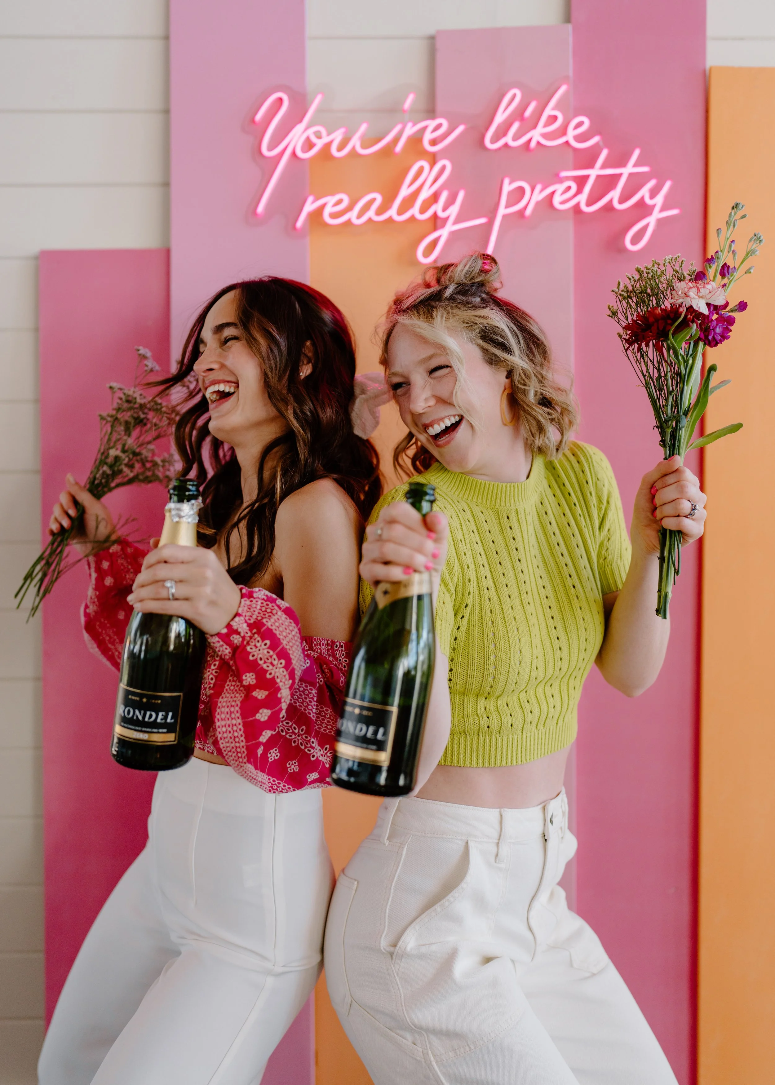 Two women celebrating with champagne bottles and flowers, smiling and enjoying themselves in front of a colorful background with a pink neon sign that reads 'You're like really pretty'.
