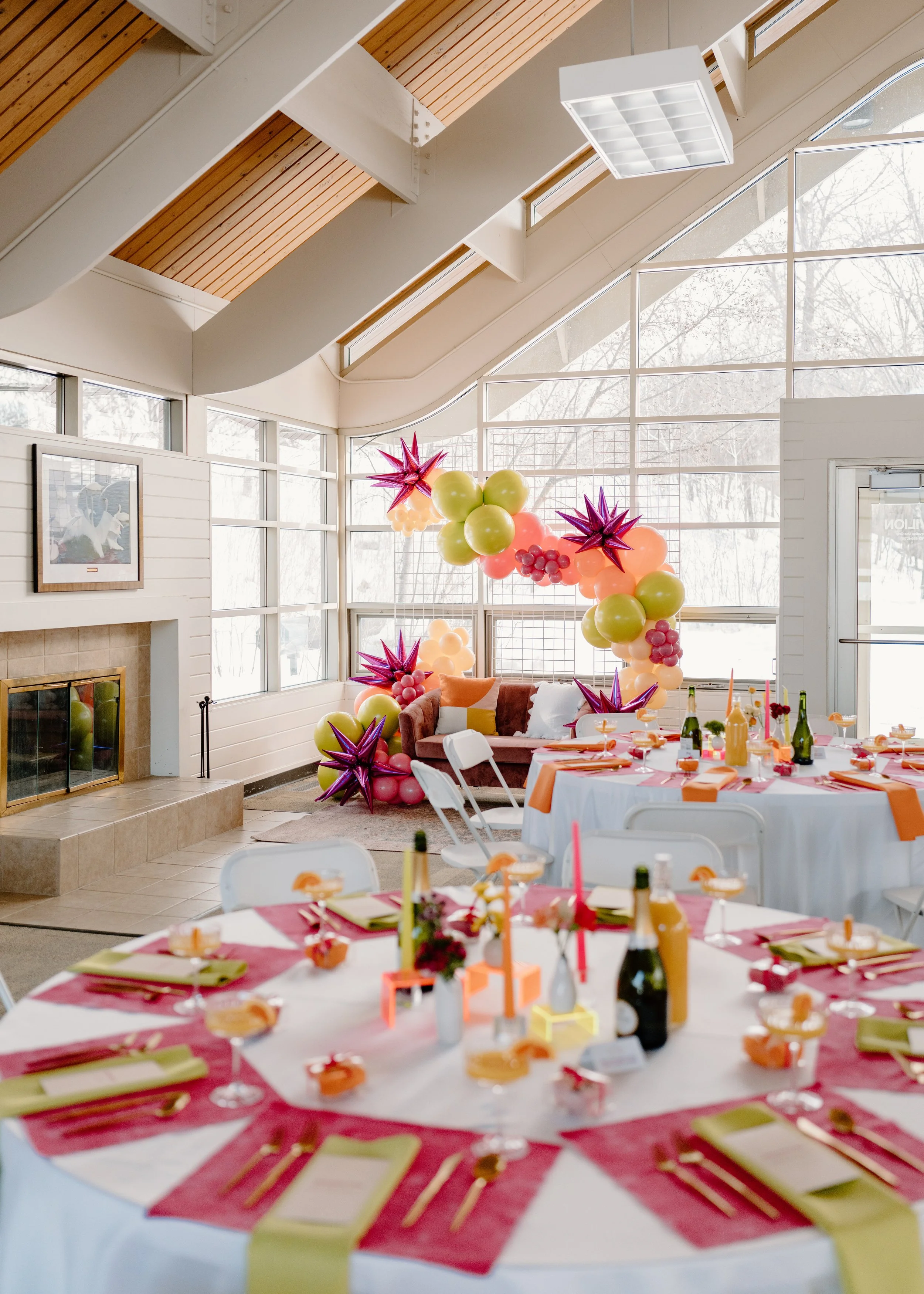 Indoor party setup with balloons, decorated tables, and a colorful balloon arch in a bright, modern room with large windows and a fireplace.