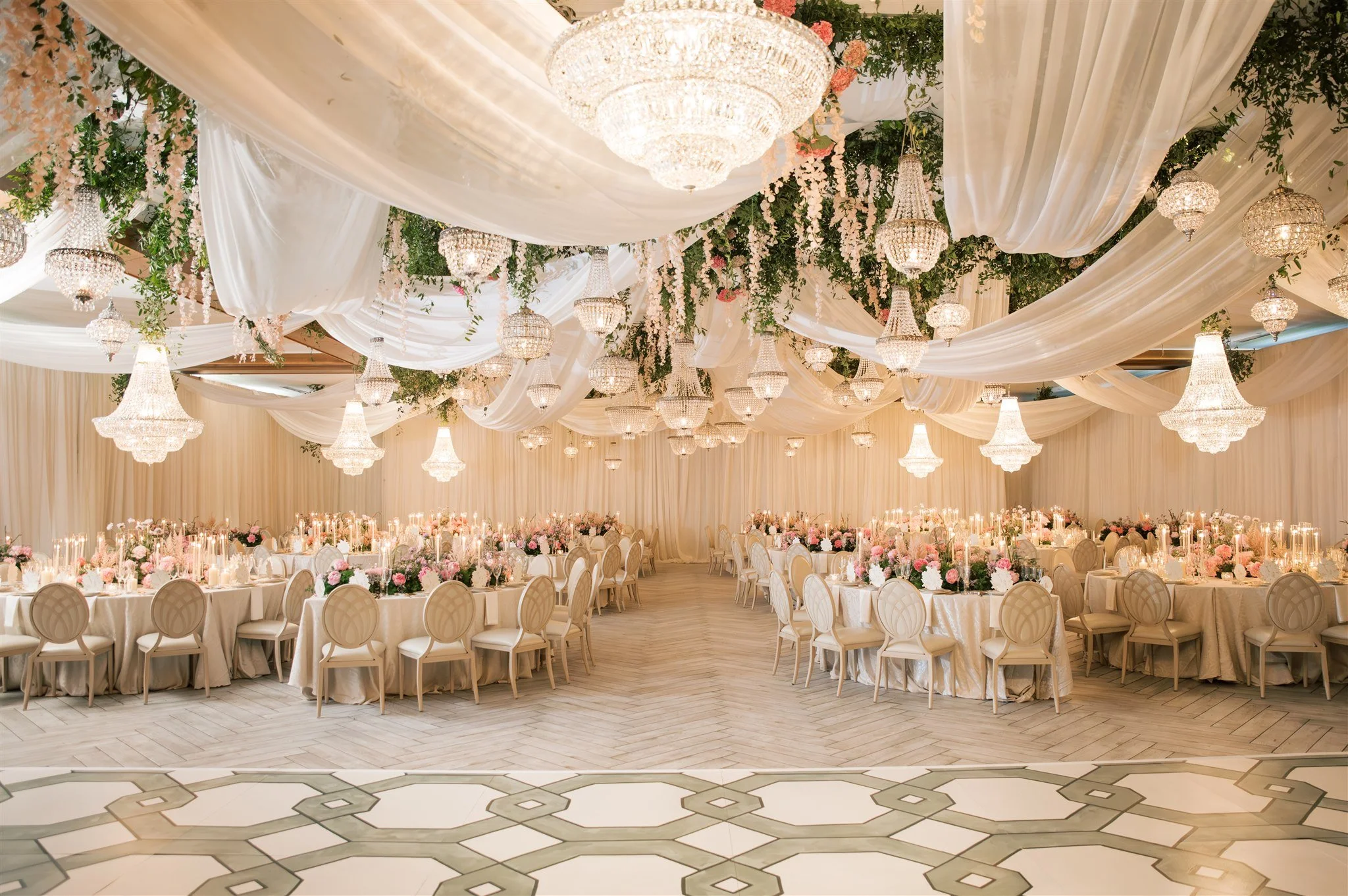 Elegant banquet hall decorated with white drapes, lush greenery, and pink floral arrangements. Multiple chandeliers hang from the ceiling, casting a warm glow over round tables set for a formal event.