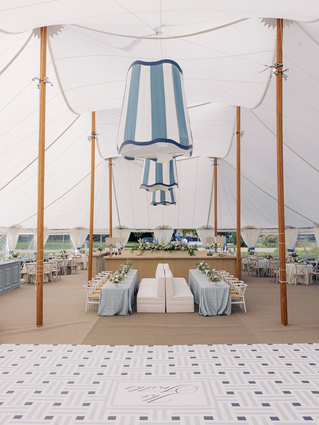 Elegant event setup under a large white tent with blue and white striped patio umbrellas hanging from the ceiling, a decorated stage with floral arrangements, and tables set with cloths and floral centerpieces for a gathering or celebration.