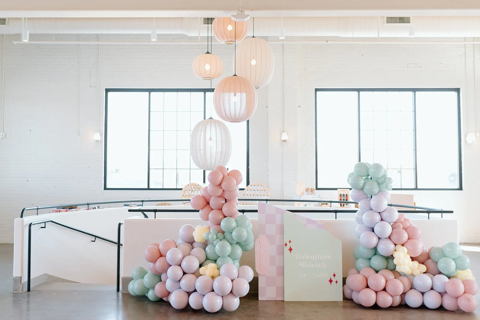Decorative balloons in pastel pink, mint green, and cream colors arranged in clusters at a shop entrance with a sign that says "Valentine's Brunch SIP SHOP," inside a bright, modern space with white brick walls, large windows, and hanging paper lantern lights.