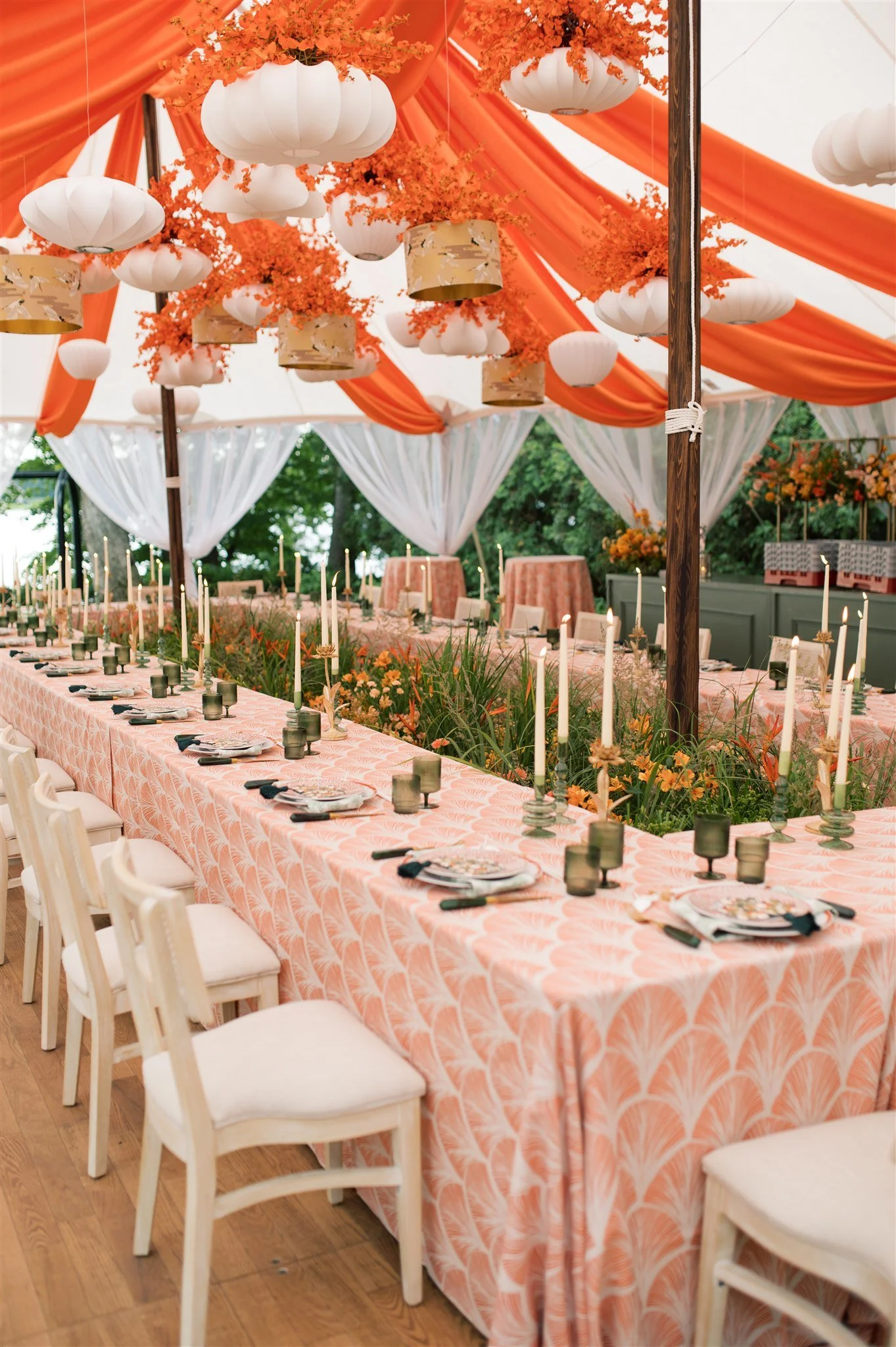 A decorated outdoor dining area with a long peach-colored tablecloth-covered table set with plates, glasses, and napkins, surrounded by chairs. The tent roof is decorated with orange and white fabric, hanging white paper lanterns, and orange foliage. Candles and greenery line the center of the table, creating an elegant setup for a celebration or event.