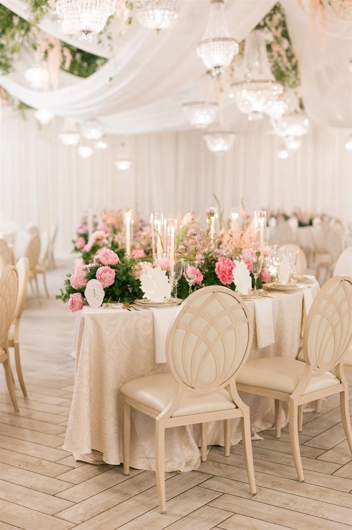 Elegant wedding reception with round table covered in cream tablecloth, adorned with pink and white floral centerpieces, candles, and fine dinnerware, set in a decorated venue with draped fabric and chandeliers.