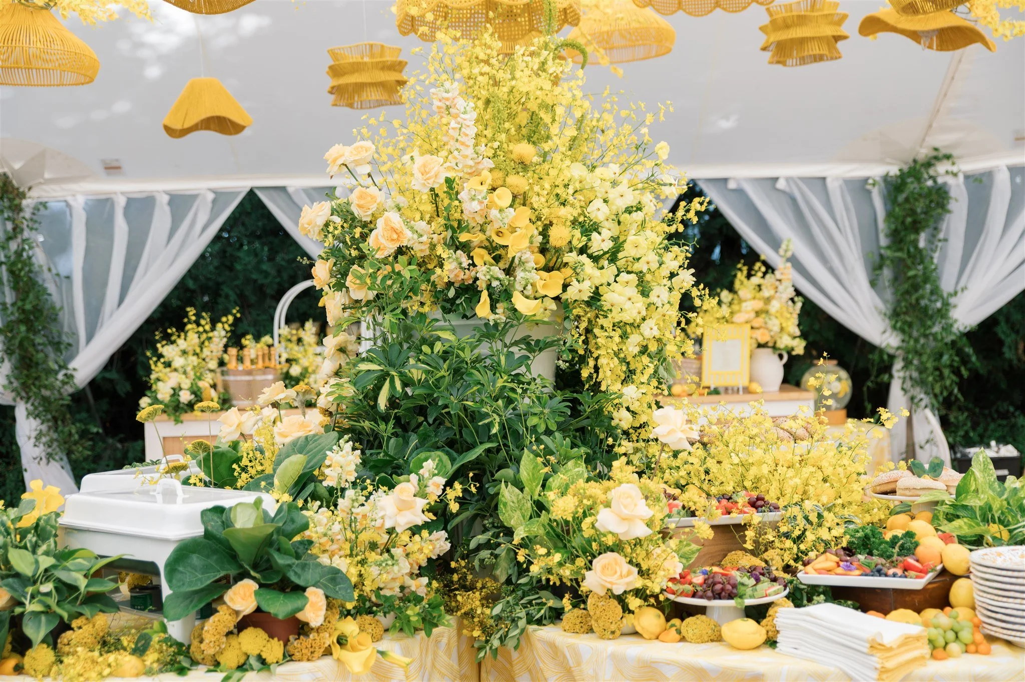Decorative outdoor table covered with yellow and white flowers, fruit, and tableware, with white draped canopy and hanging yellow lanterns.