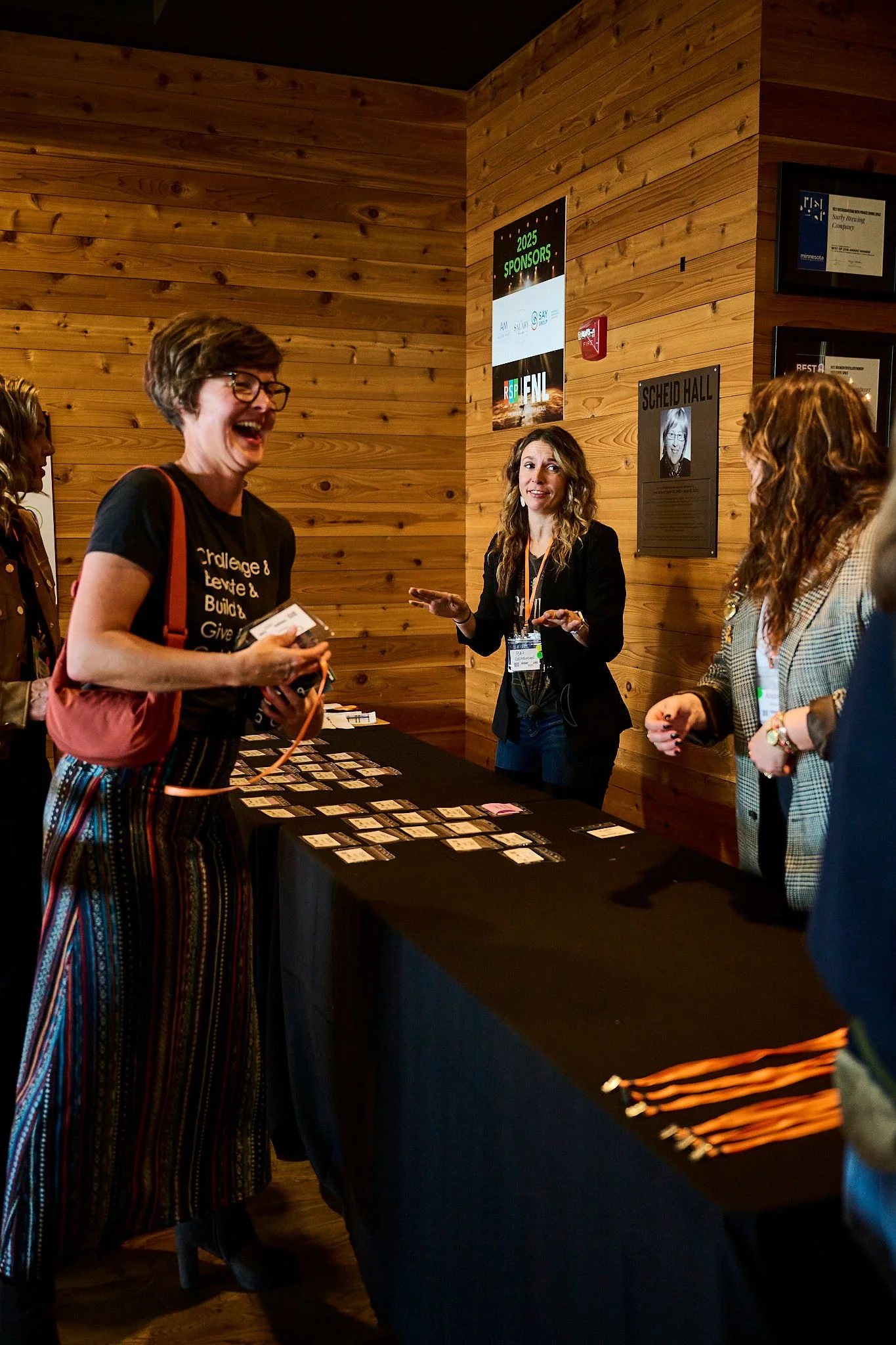 People standing around a table with name tags, engaging in conversation at an indoor event with wooden walls.