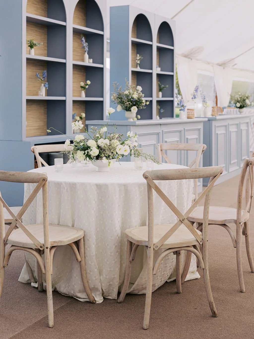 A round table with a white lace tablecloth, decorated with a floral centerpiece of white flowers and greenery, surrounded by light wood chairs, set inside a light blue and white decorated event space with built-in shelves and additional floral arrangements in vases.