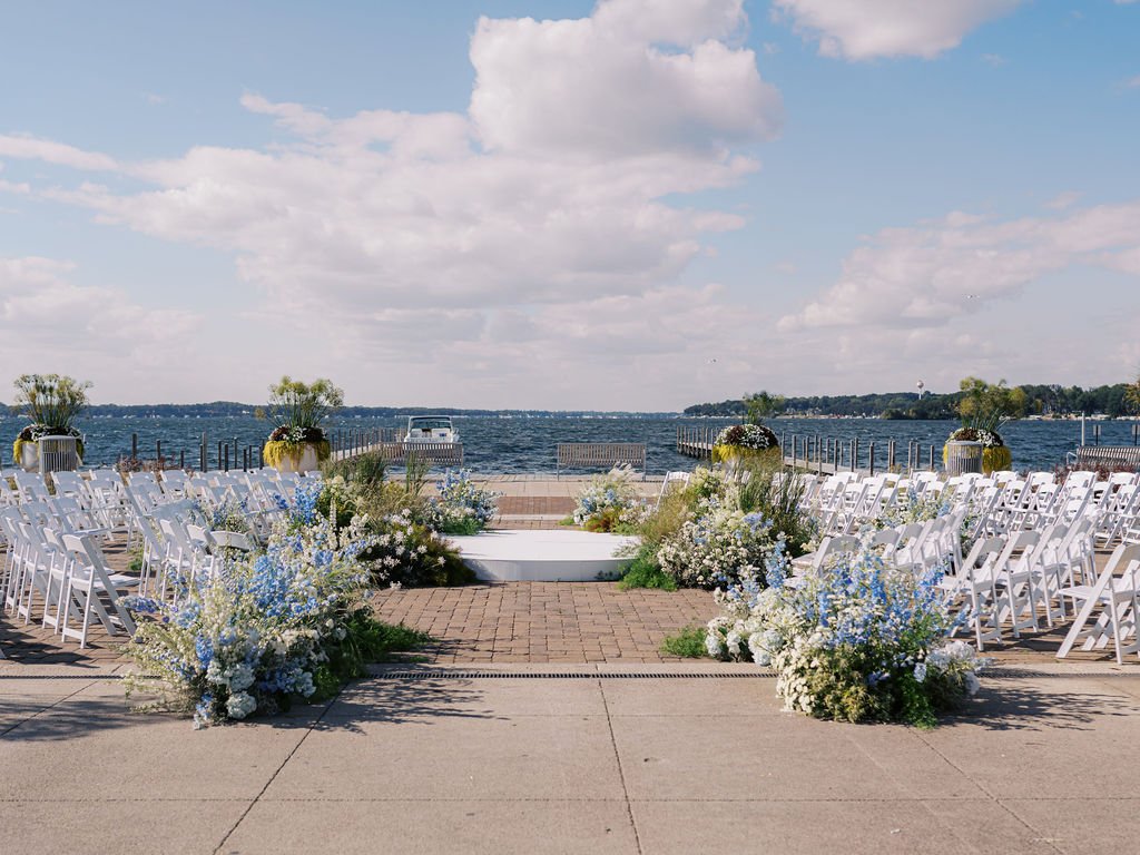 Outdoor wedding ceremony setup on a dock with white chairs, floral arrangements, and a view of a lake under a partly cloudy sky.