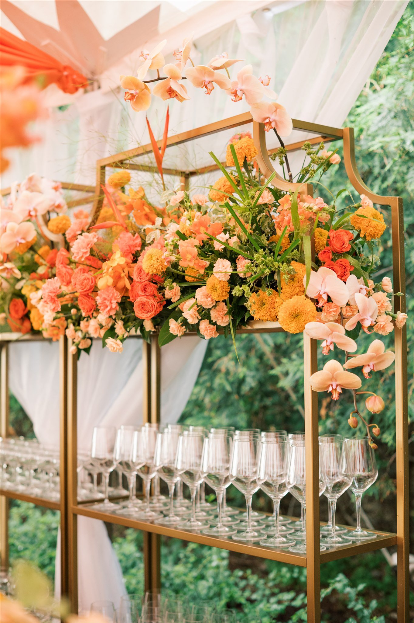 A decorative display of pink and orange flowers arranged on a gold metal shelf, with empty wine glasses below, set outdoors with a white fabric backdrop and green foliage in the background.