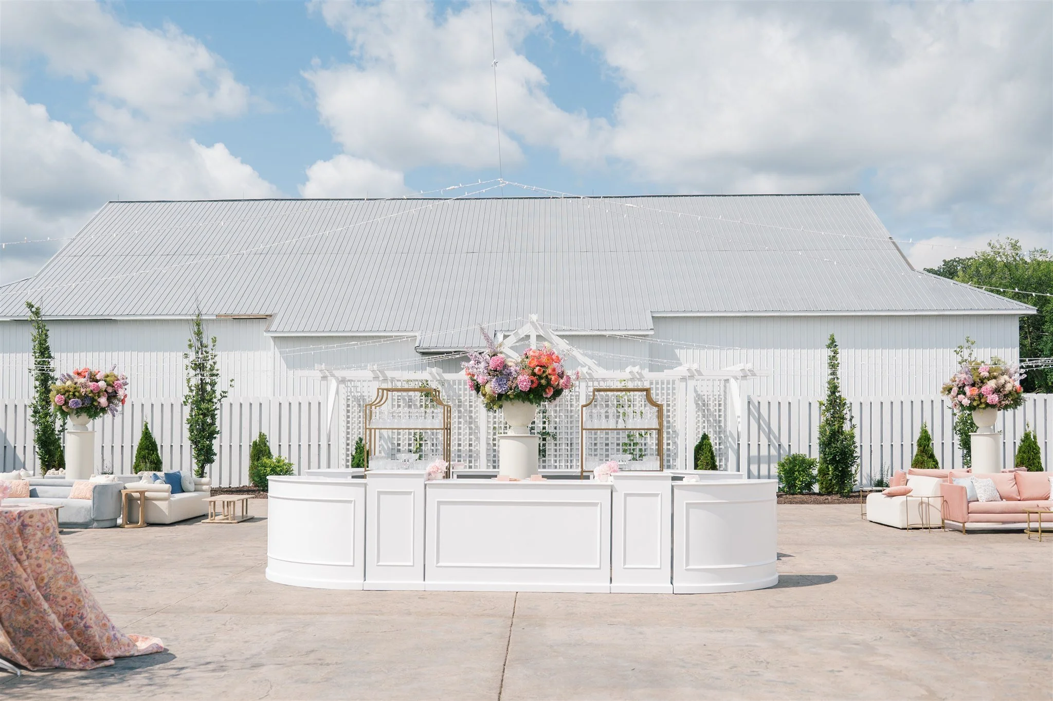 Outdoor wedding or event setup with a white arched fencing, floral arrangements, and seating, set against a barn with a metallic roof. The sky is partly cloudy.