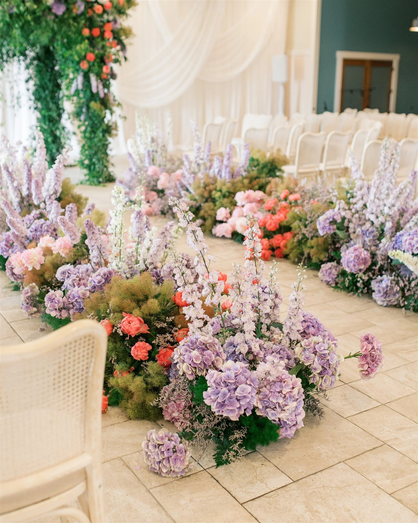 Indoor wedding aisle decorated with pastel purple, pink, and white flowers, with chairs arranged on each side and a floral archway in the background.