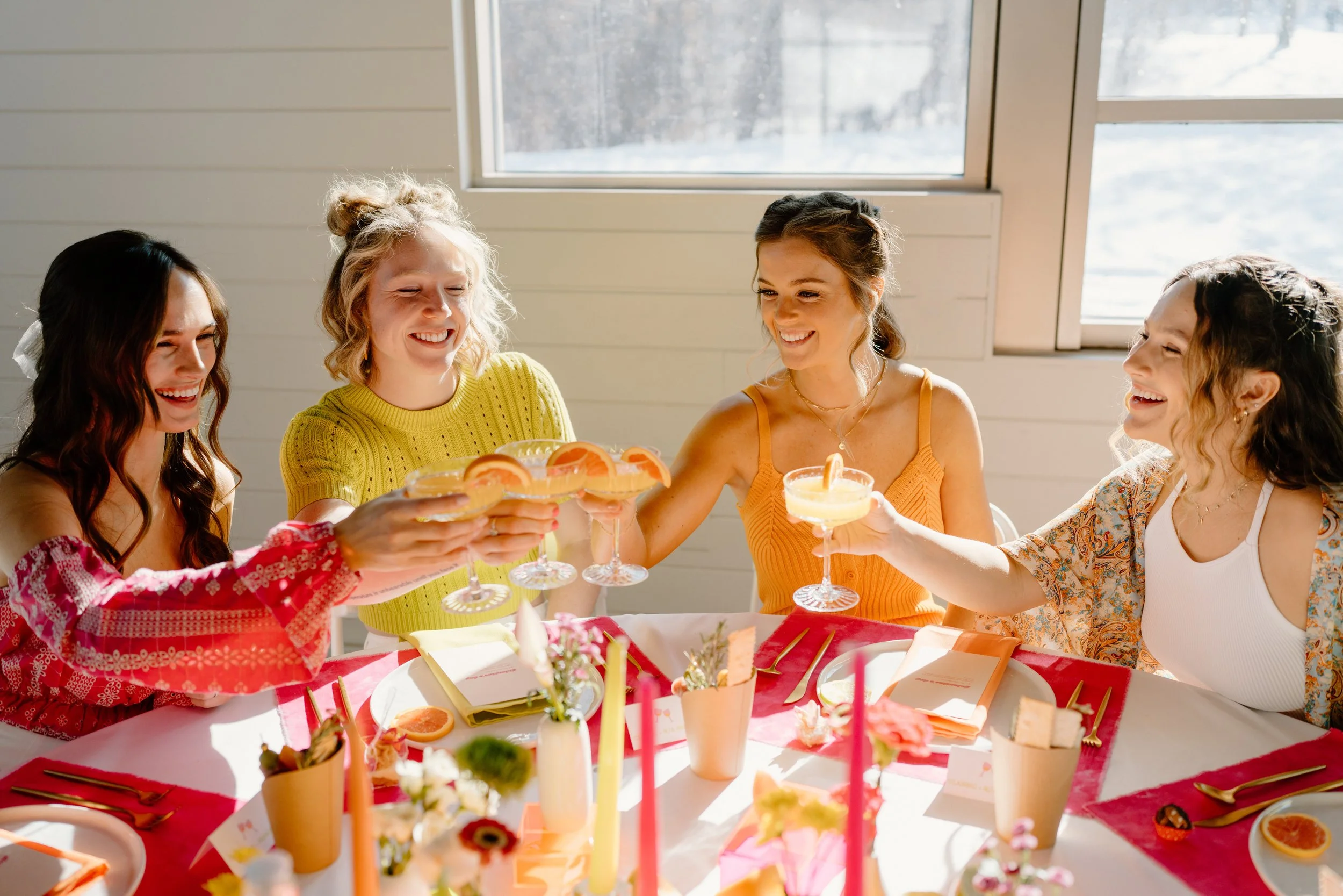 Four women smiling and raising glasses with drinks in a celebration, sitting around a decorated table with flowers and orange slices, by a window.