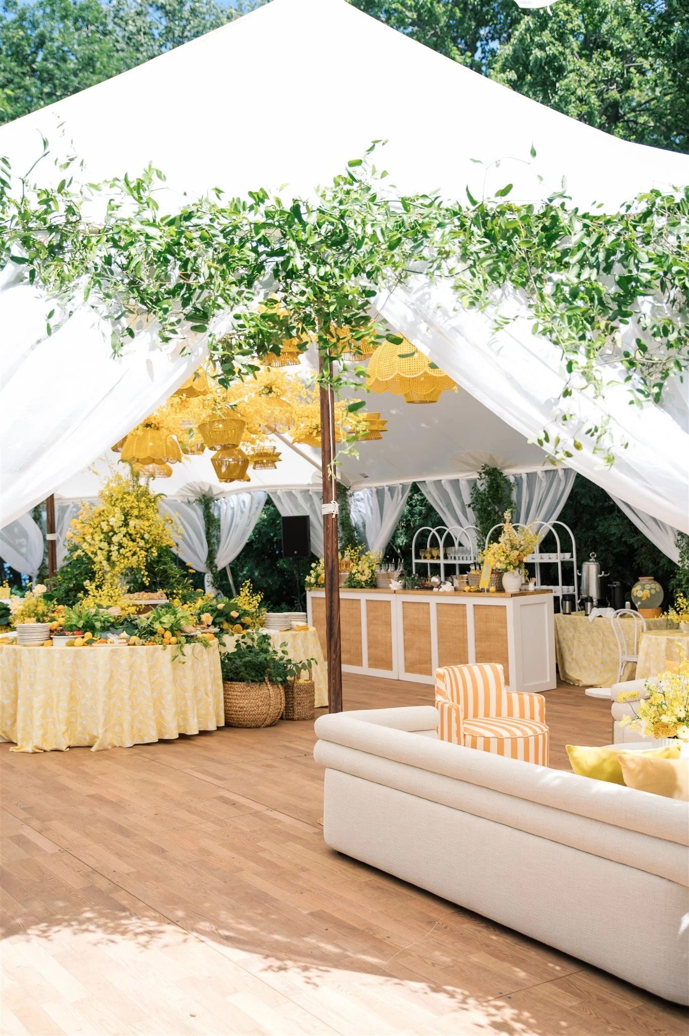 Decorated outdoor event tent with yellow-themed decor, white curtains, floral arrangements, a buffet table, and a seating area with white and striped chairs on wooden flooring.
