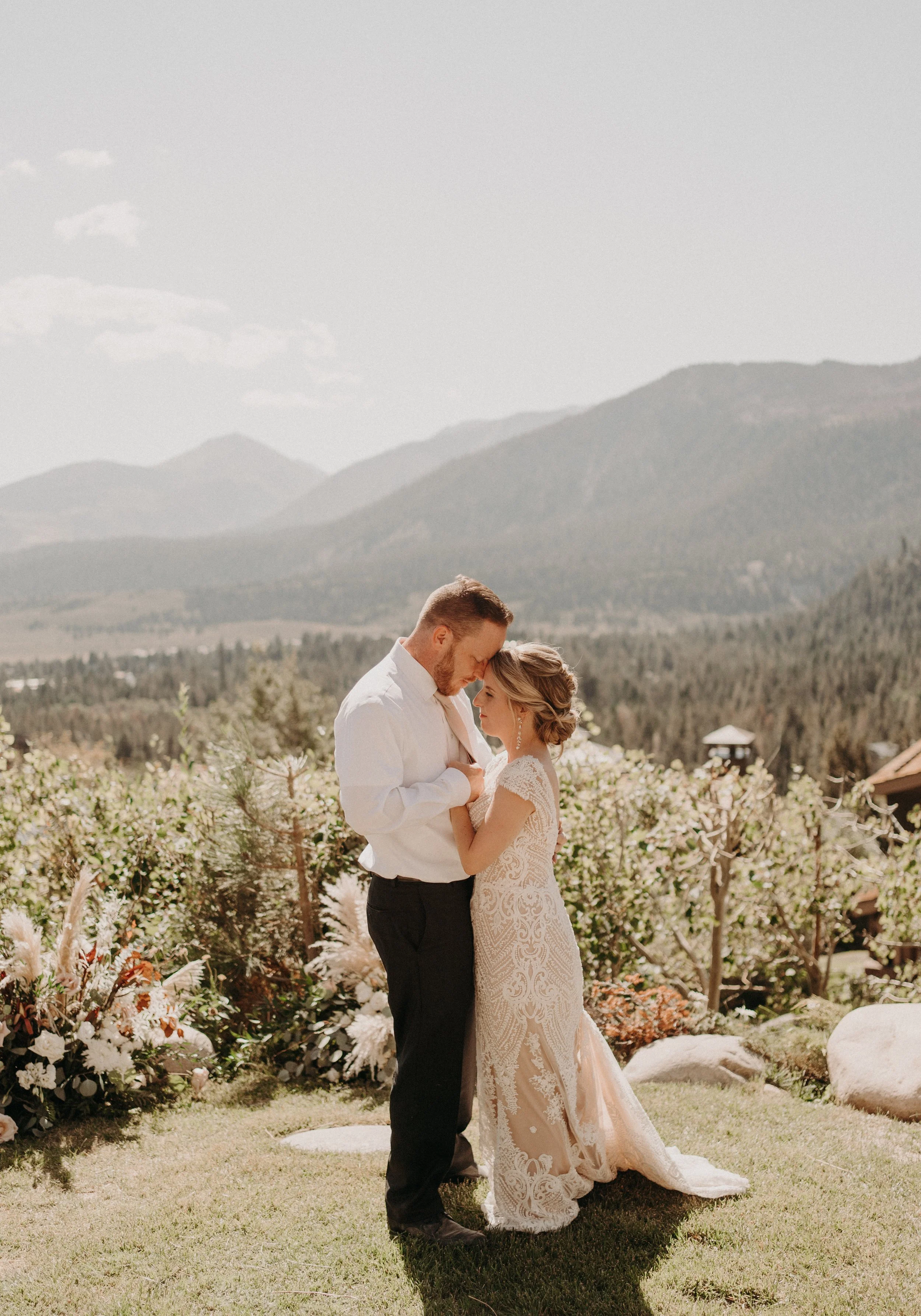A couple in wedding attire embracing outdoors with mountains and flowers in the background.