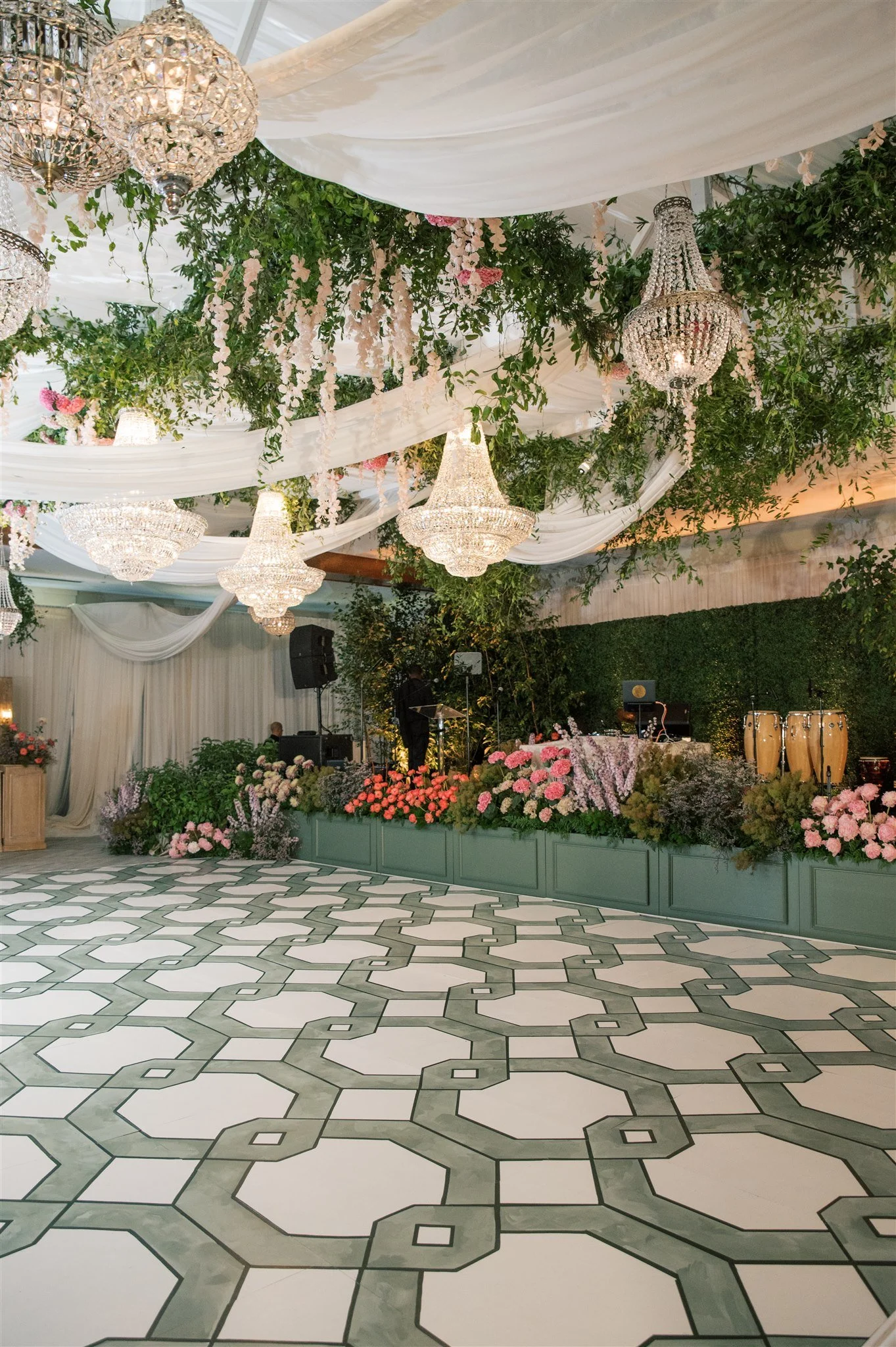 Elegant event venue decorated with chandeliers, lush greenery, and pink flowers. A stage with musical instruments is visible, and the floor has a black and white geometric tile pattern.