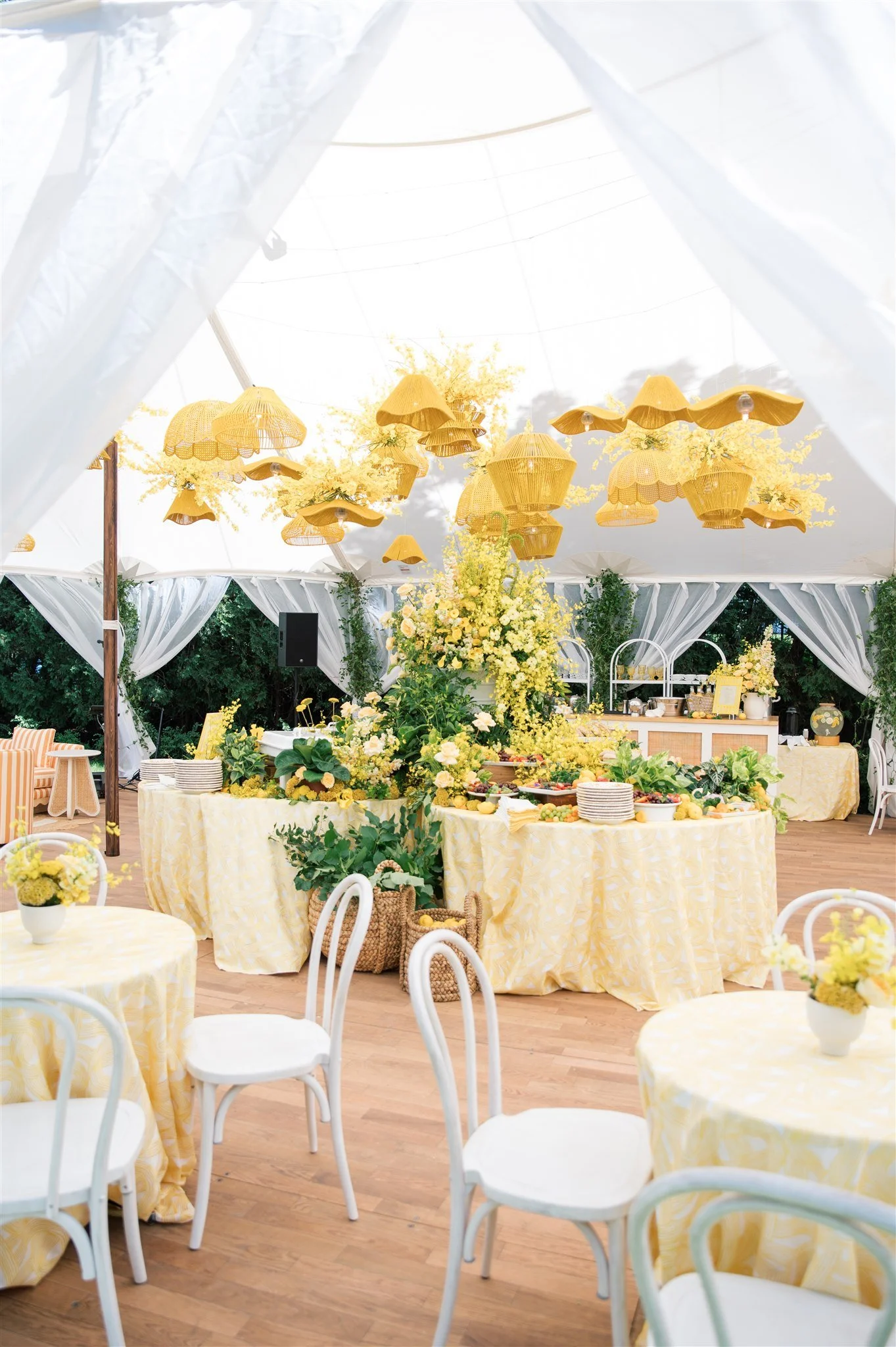 A decorated event space with yellow and white decorations, featuring yellow umbrellas and floral arrangements, with side tables and chairs, and a buffet area in the background.