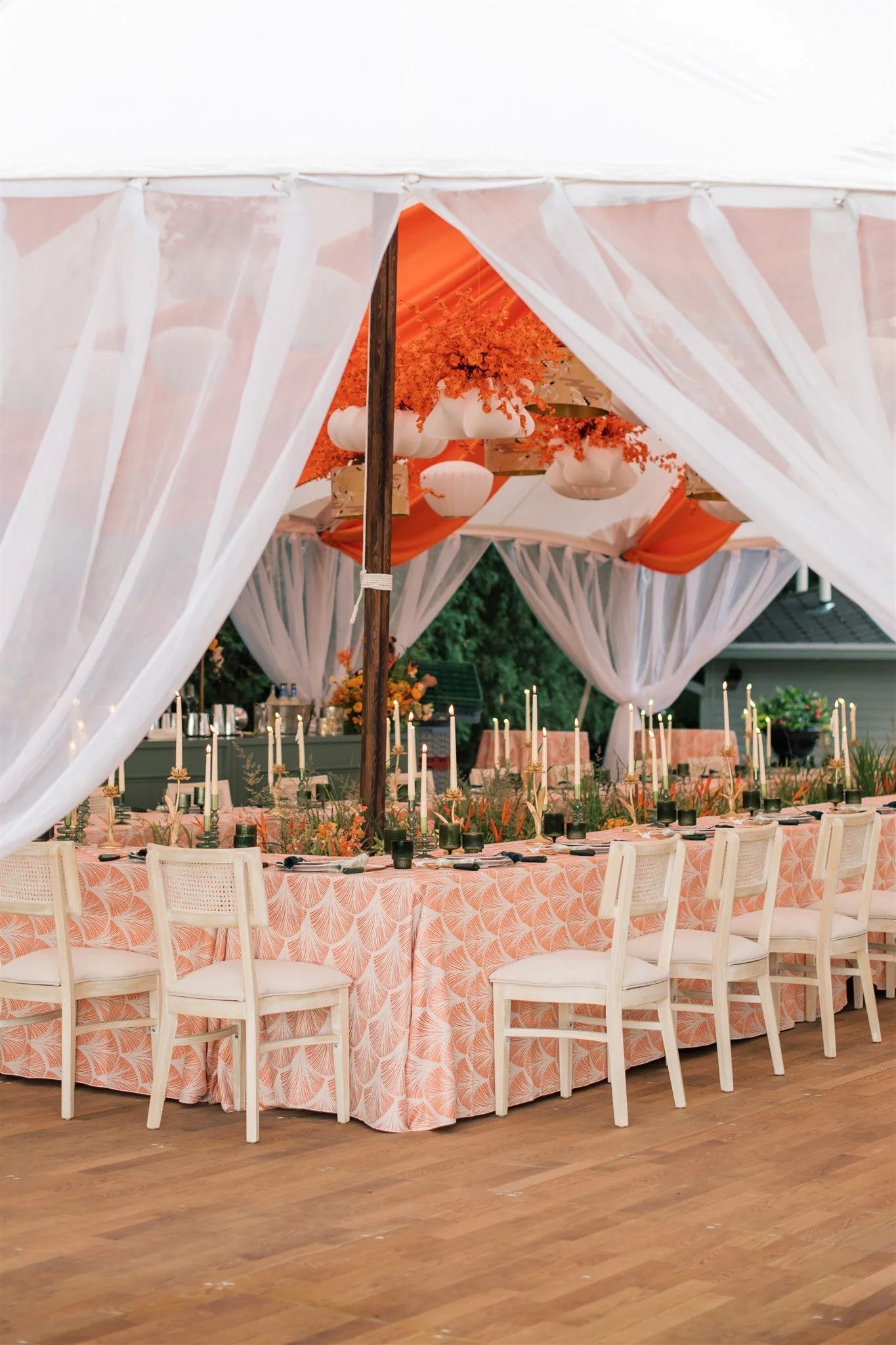 Decorative event table setup inside a large white tent with white drapes, orange accents, pink patterned tablecloth, candles, and chairs.