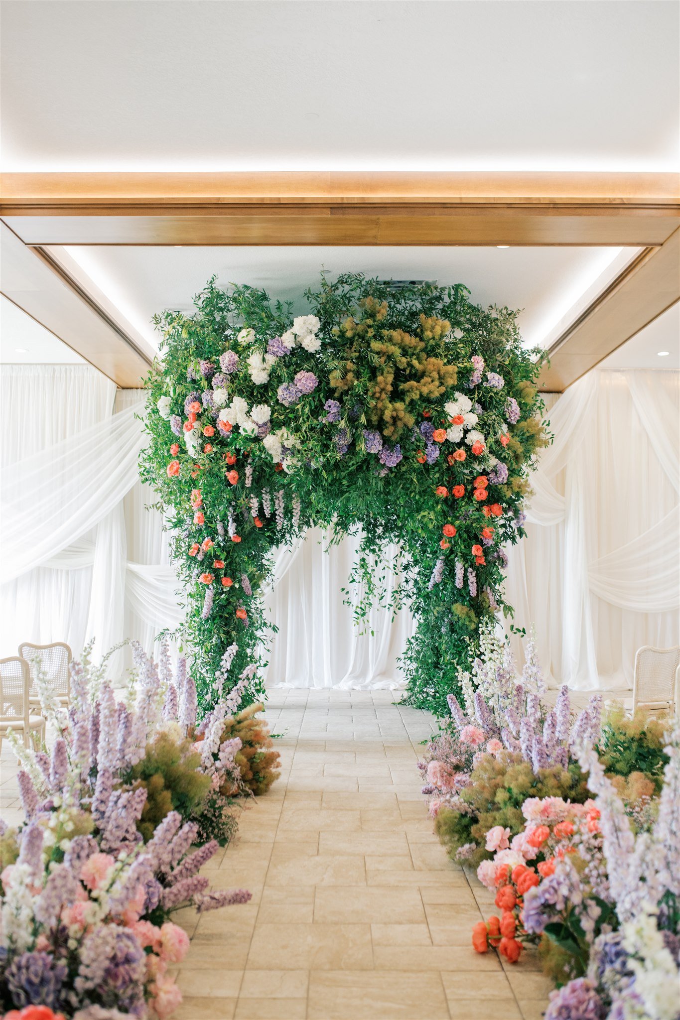 A floral wedding arch decorated with purple, white, pink, and orange flowers, with side arrangements and draped white curtains in the background.