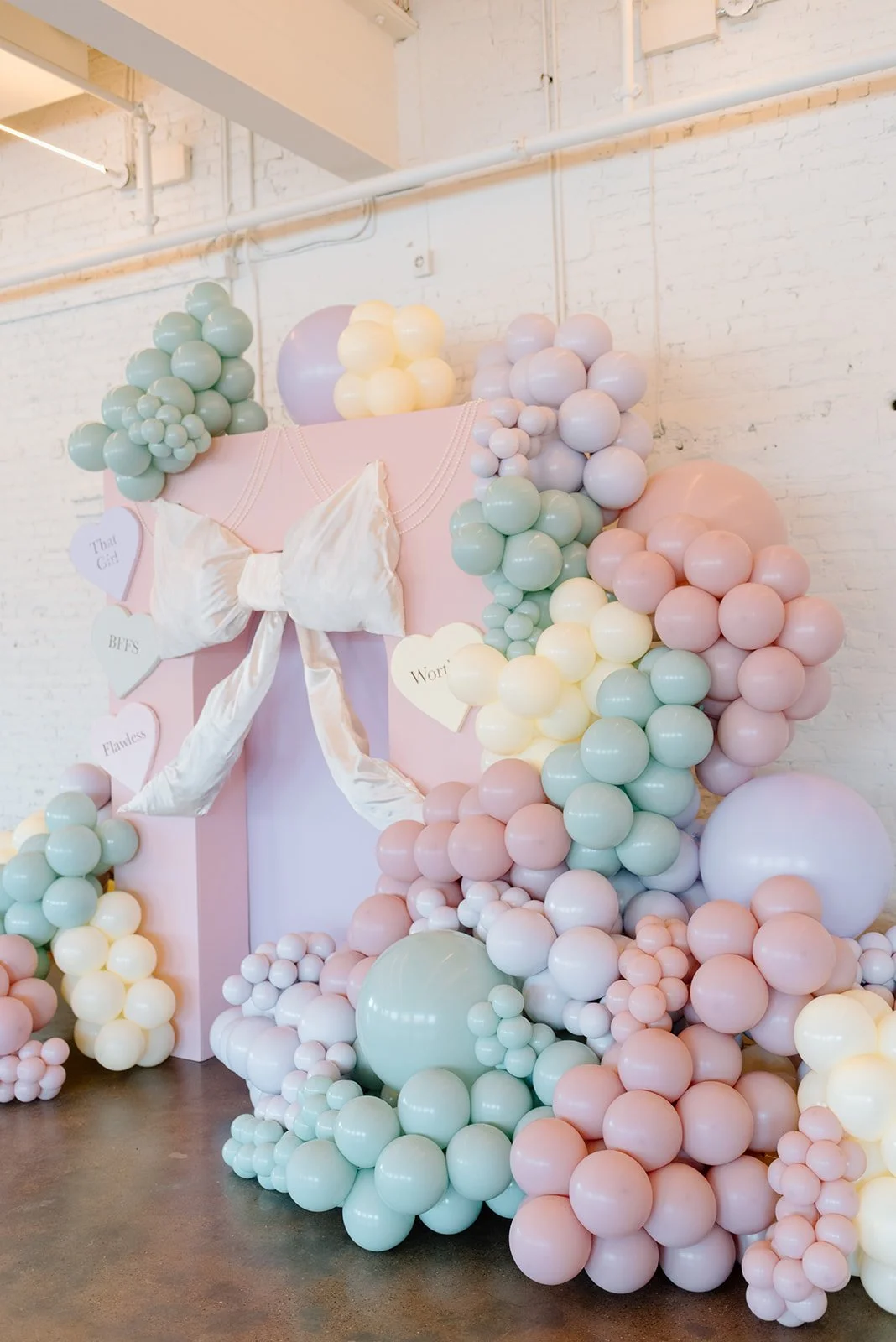Decorated wall with pastel balloons in pink, mint green, cream, and purple, arranged around a pink backdrop with a white bow and heart-shaped signs.