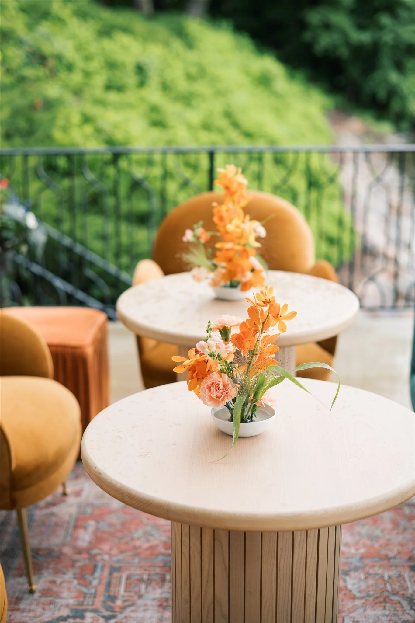 Decorative orange and pink floral arrangements on white round tables on an outdoor patio with orange and yellow chairs and a green lush background.