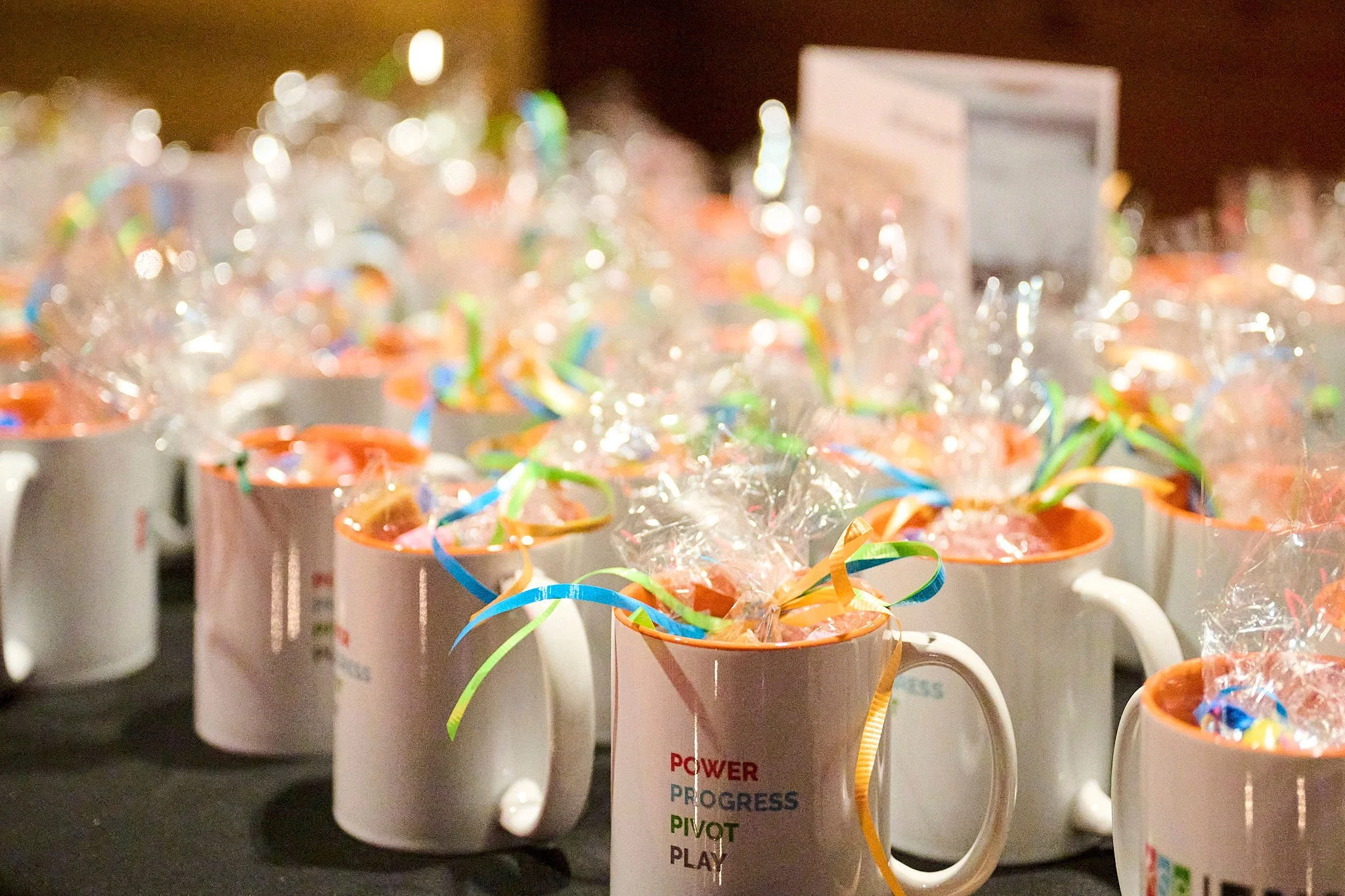 Multiple white coffee mugs filled with candy, decorated with colorful ribbons and clear plastic wrapping, placed on a dark surface.