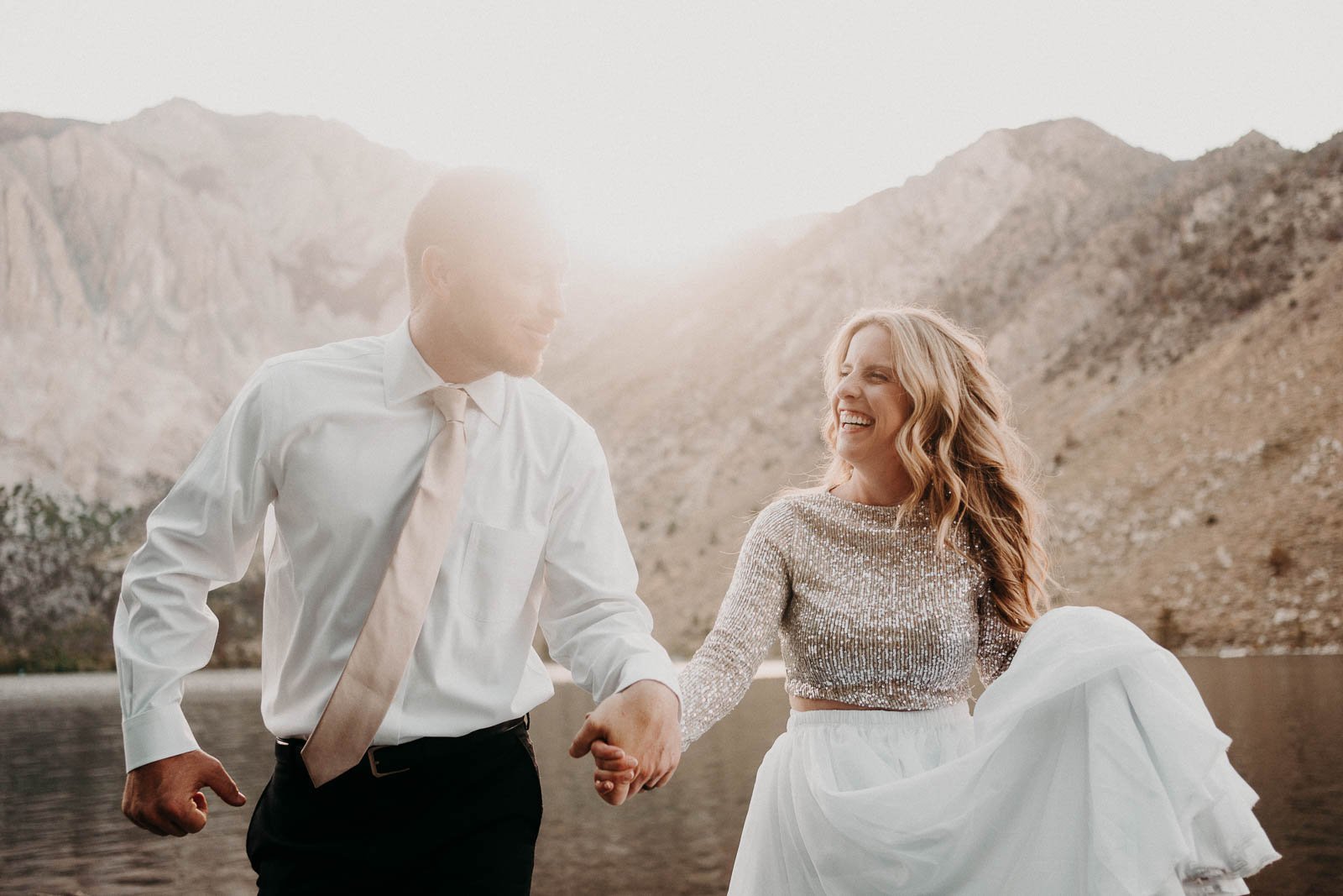 A smiling woman in a white dress holding a white garment and a man in a white shirt and tie holding hands, walking outdoors near water with mountains in the background at sunset.