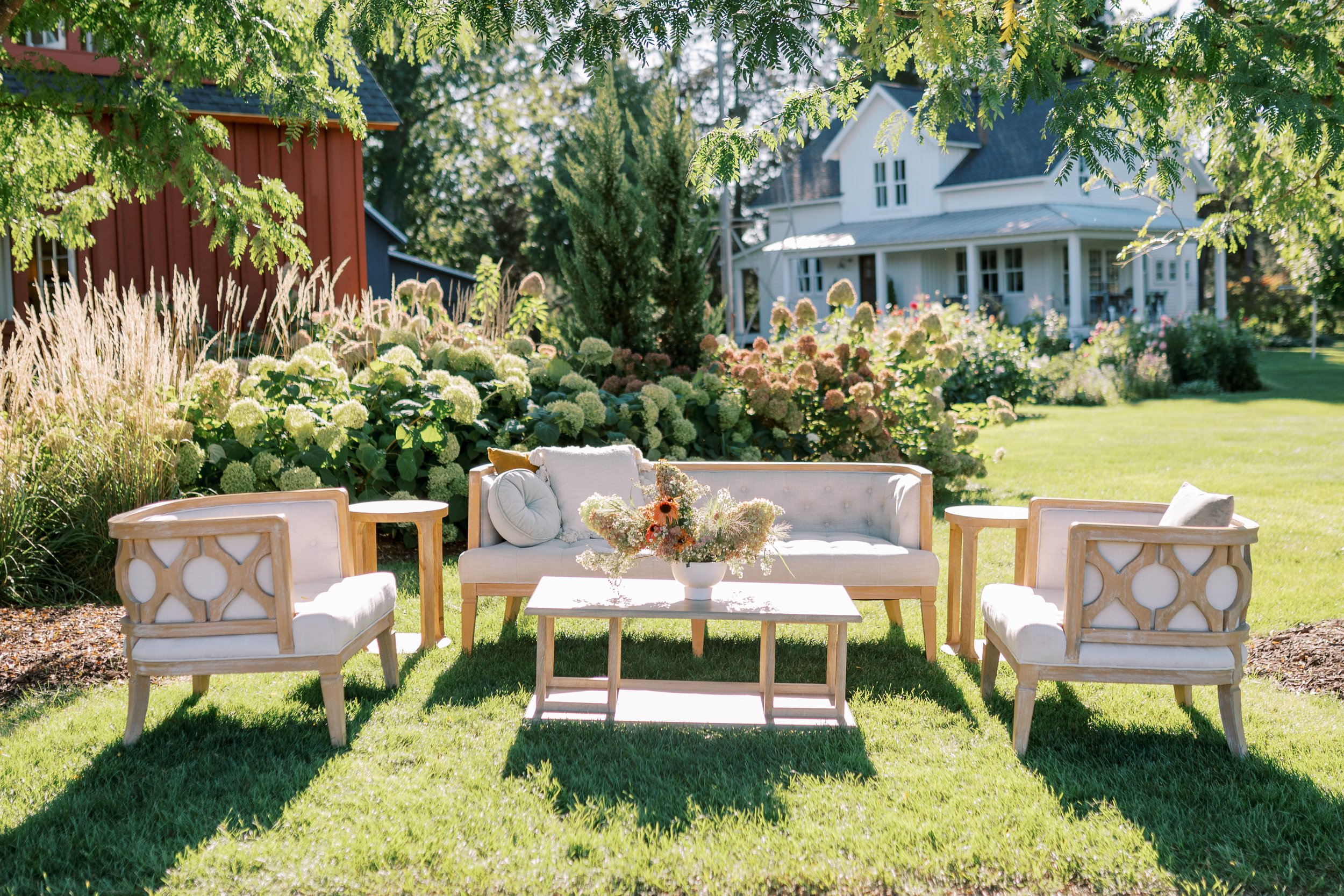Outdoor garden seating area with a white sofa and two matching chairs around a white coffee table with a floral arrangement. Lush green grass and colorful flowering bushes surround the area, with a white house and trees in the background.