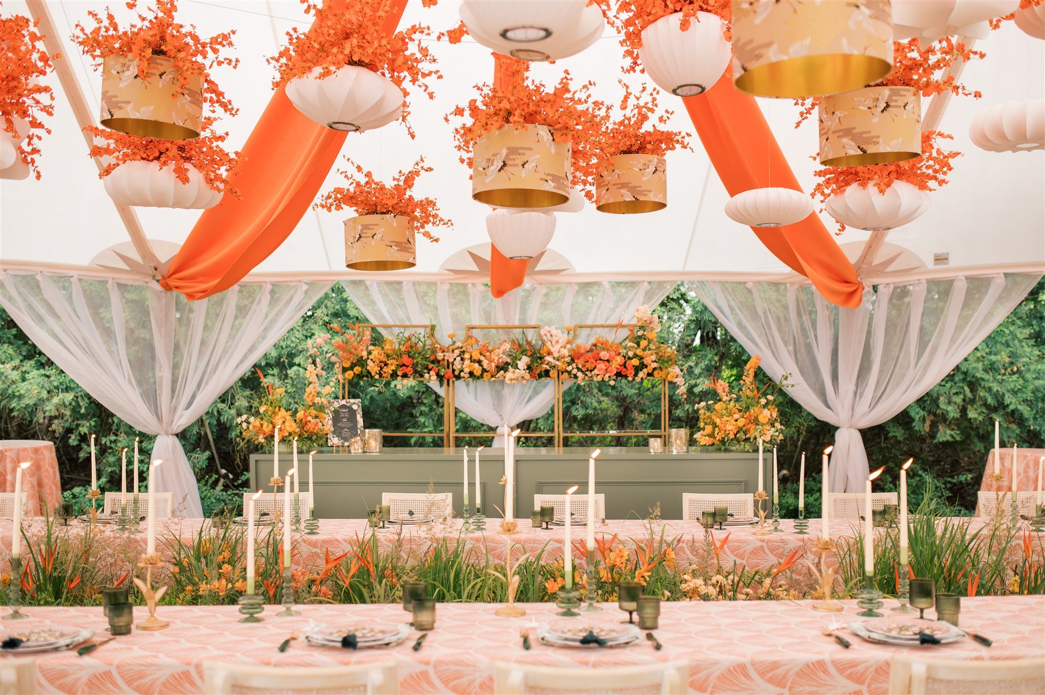 Elegant outdoor event tent decorated with orange drapes, white lanterns with floral accents, and hanging orange flowers. Long tables with pink patterned tablecloths are set with black plates, candles, and black cups, surrounded by white chairs. The background shows lush green trees.