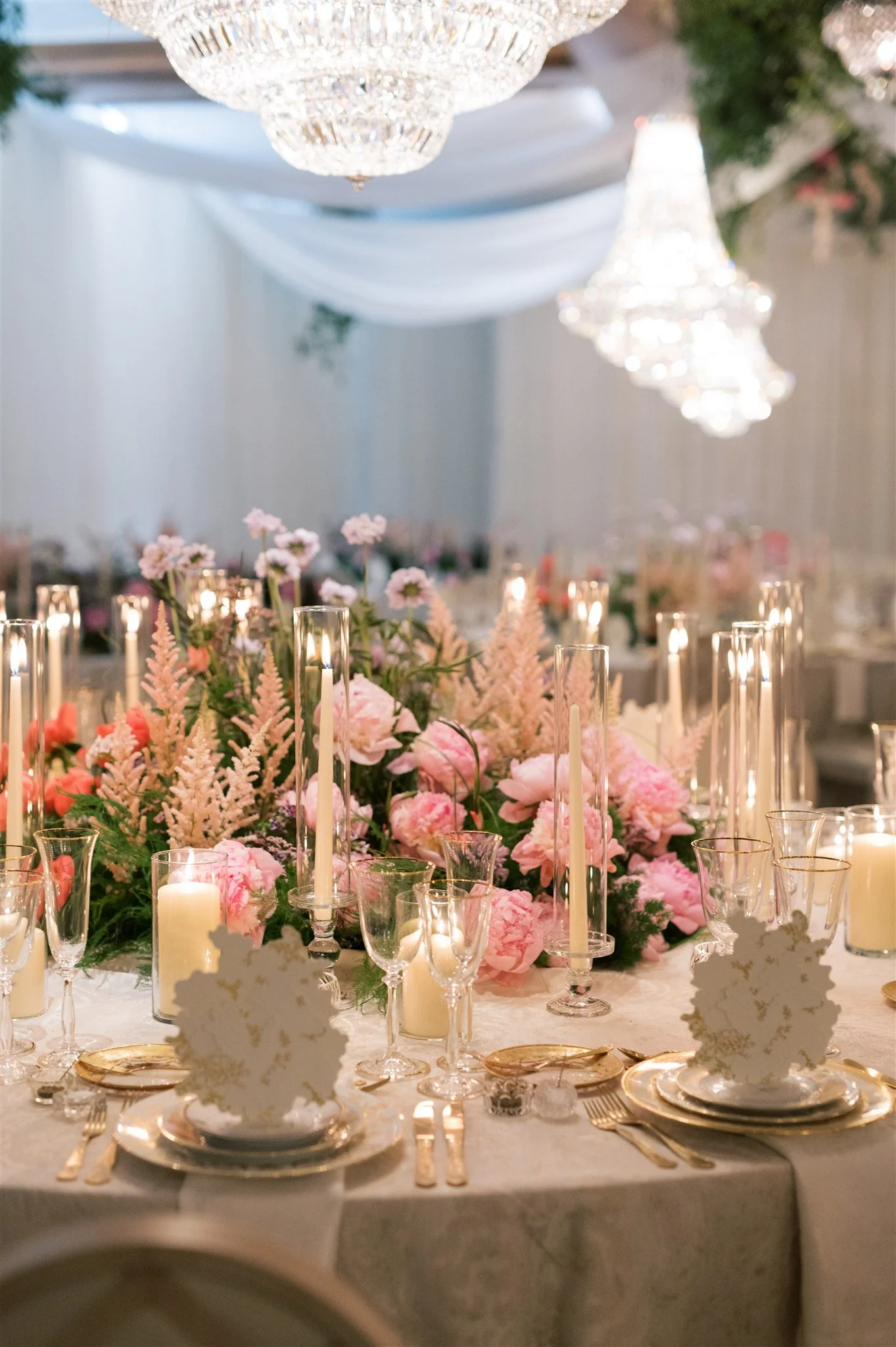 Elegant wedding or event table decorated with pink flowers, candles in glass holders, and gold-rimmed plates and utensils, illuminated by chandeliers.