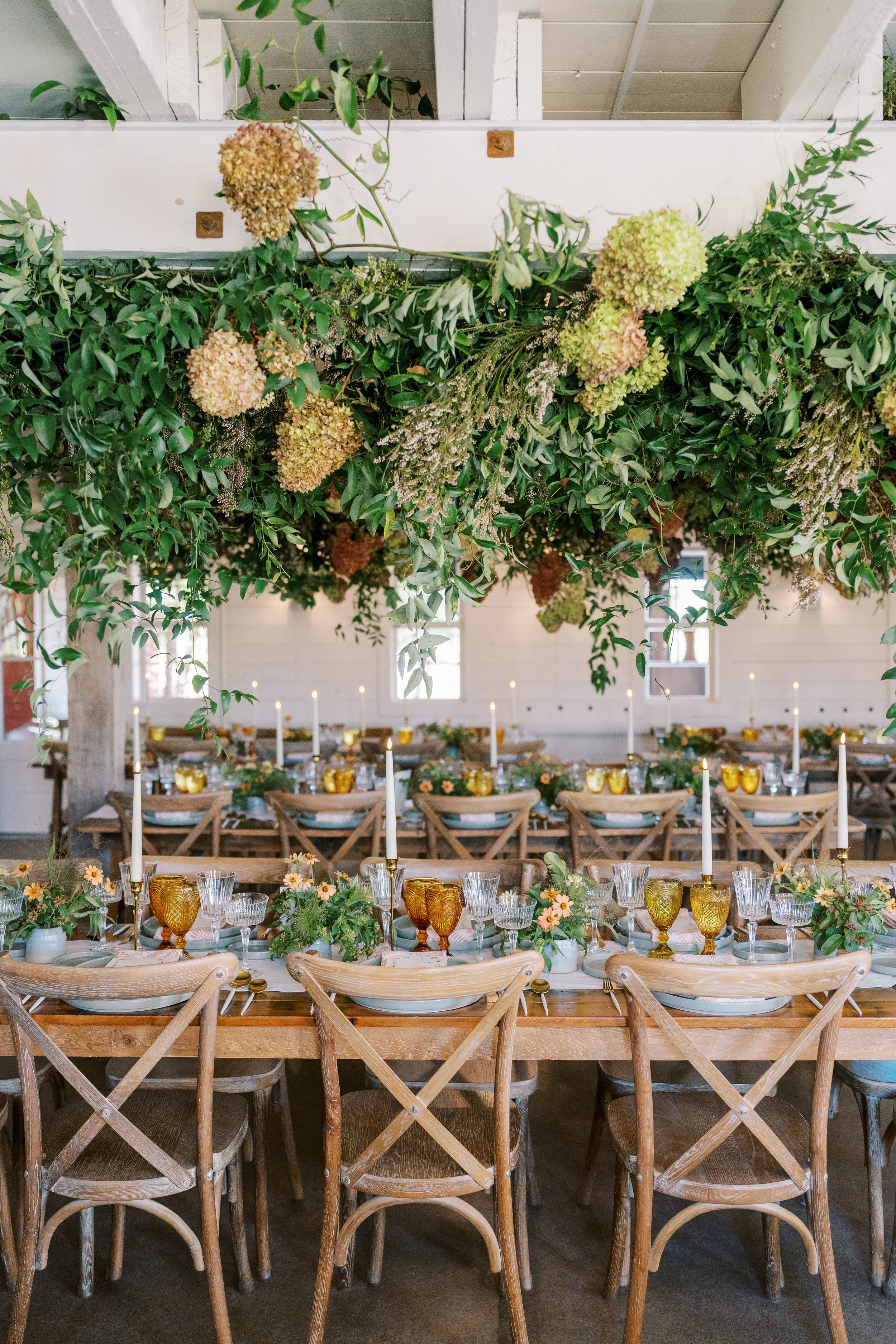 Decorated tables with candles, glassware, and floral centerpieces in a rustic setting with a hanging floral arrangement overhead.