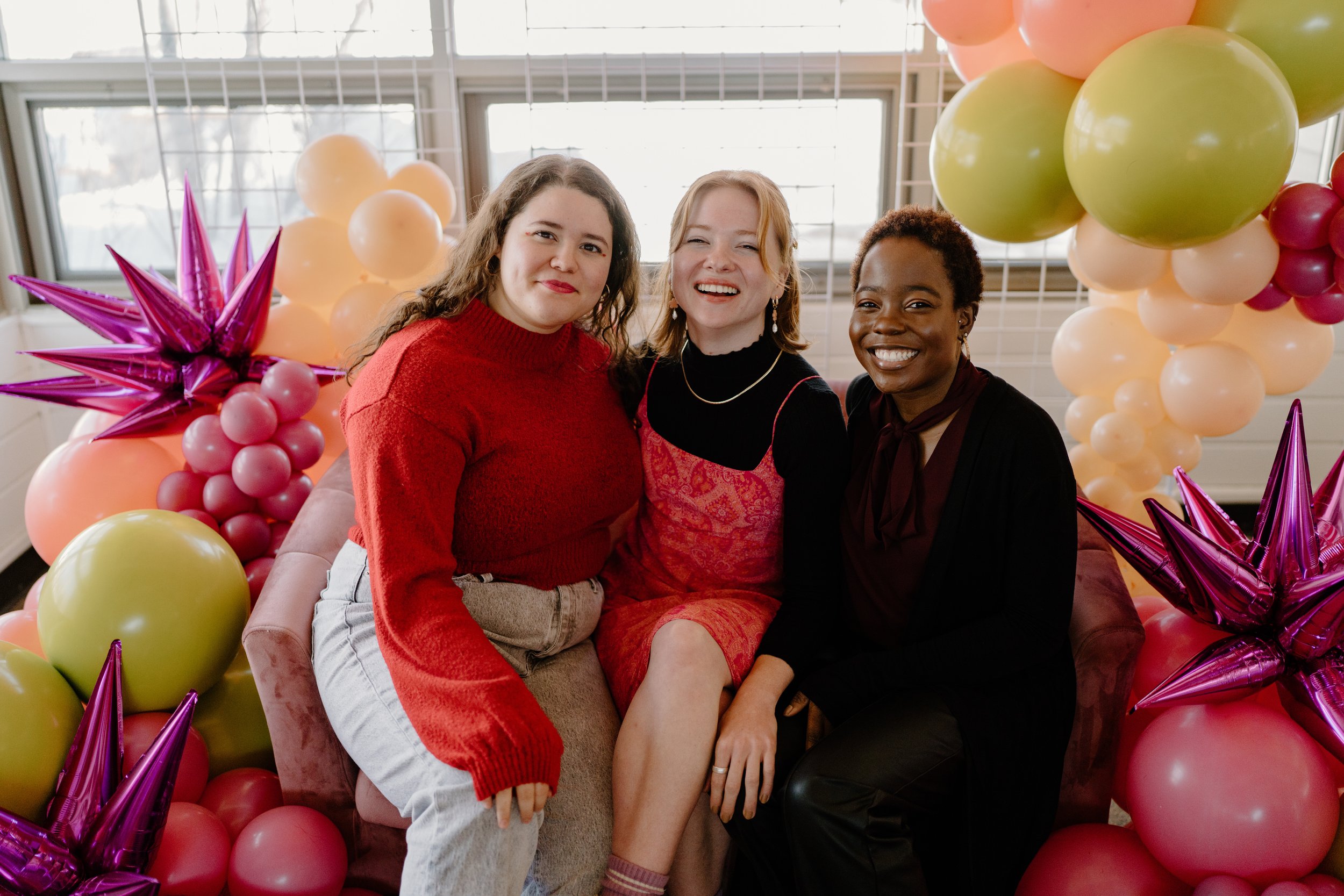 Three women sitting on a pink couch surrounded by colorful balloons, smiling at the camera.