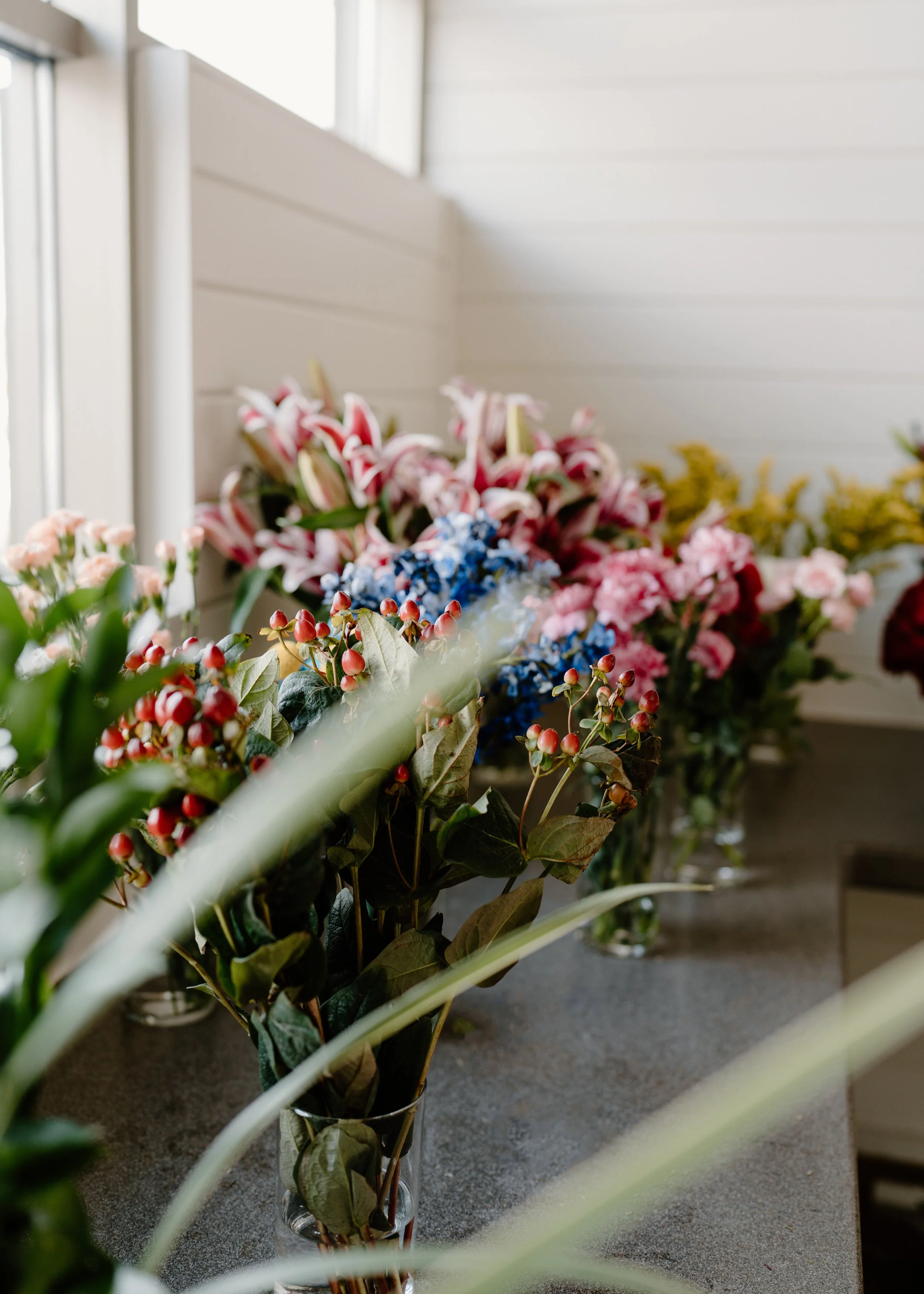 Several vases of mixed colorful flowers, including lilies, roses, and berries, on a gray tabletop in front of a white-paneled wall.