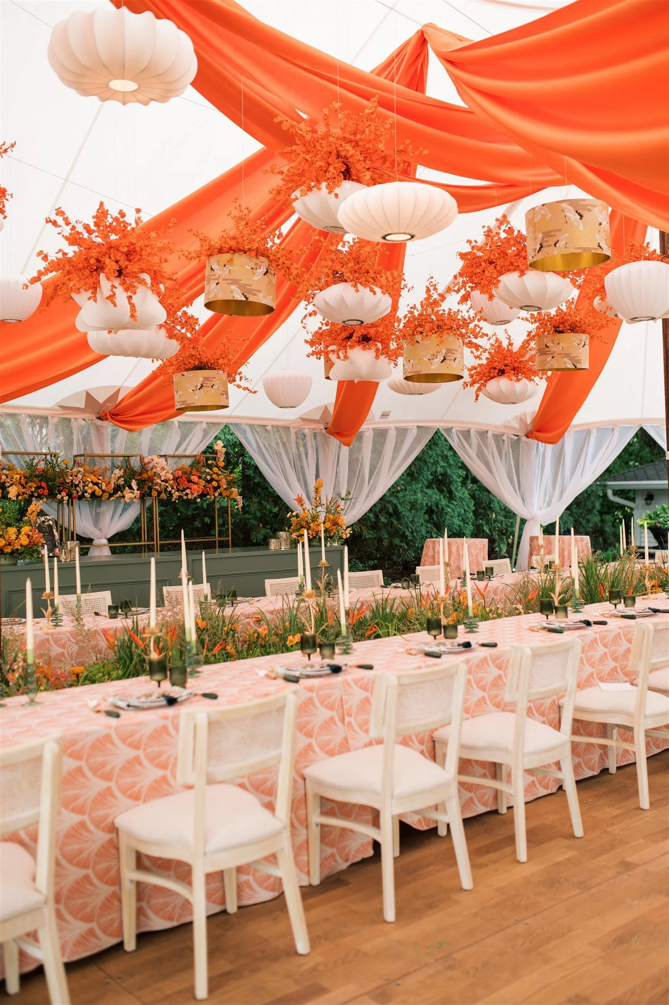 Decorated outdoor banquet table with peach tablecloth, white chairs, black glasses, candles, and floral centerpieces, under a tent with orange fabric, hanging lanterns, and orange and white floral arrangements.