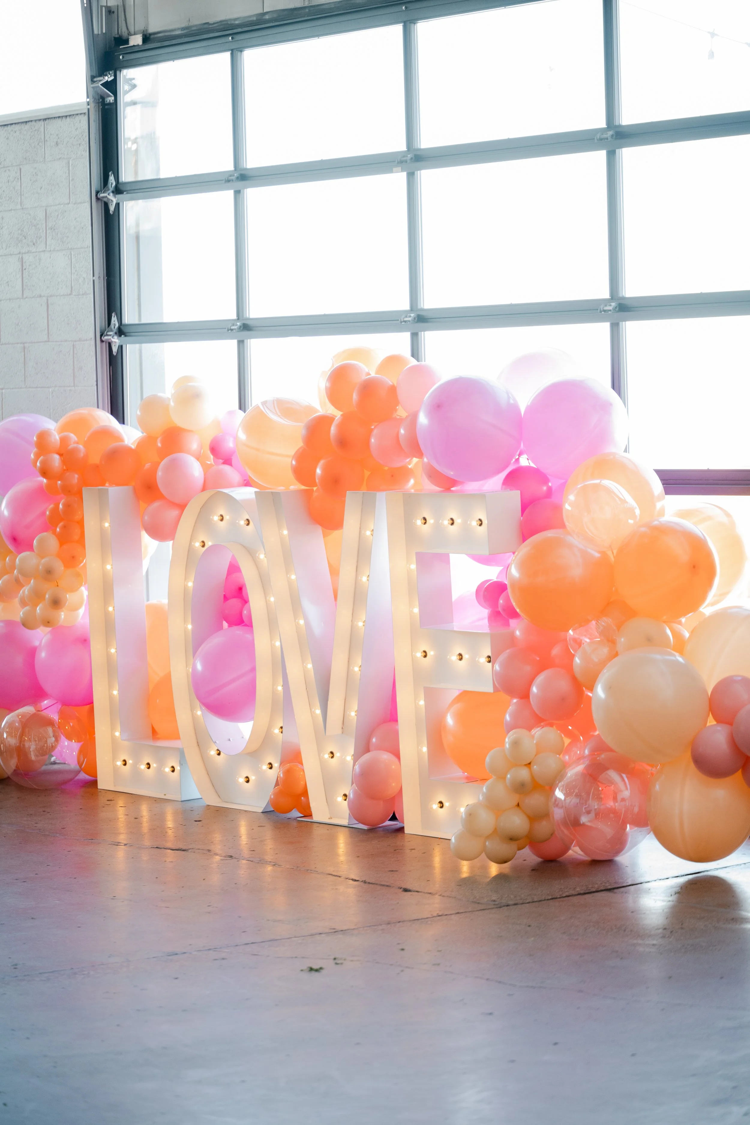 Decorative display with large illuminated letters spelling 'LOVE' surrounded by pink, orange, and peach balloons in a bright indoor setting with large windows.